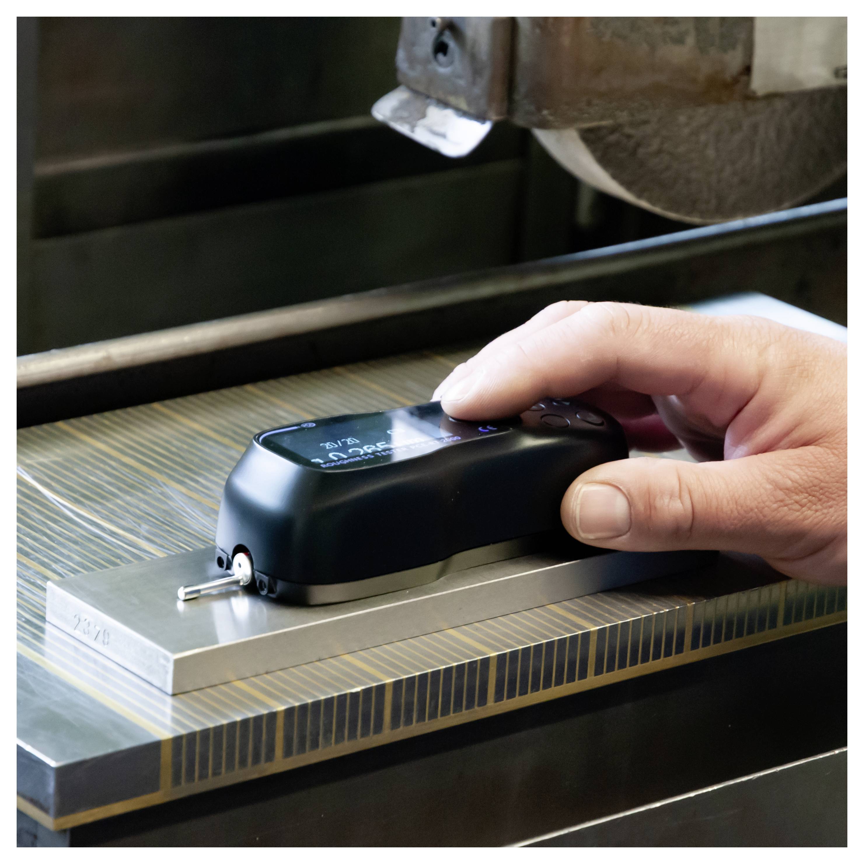 Hand hält ein schwarzes digitales Messinstrument auf einer Metalloberfläche unter einem Bearbeitungswerkzeug, um ein Teil zu messen oder zu kalibrieren.