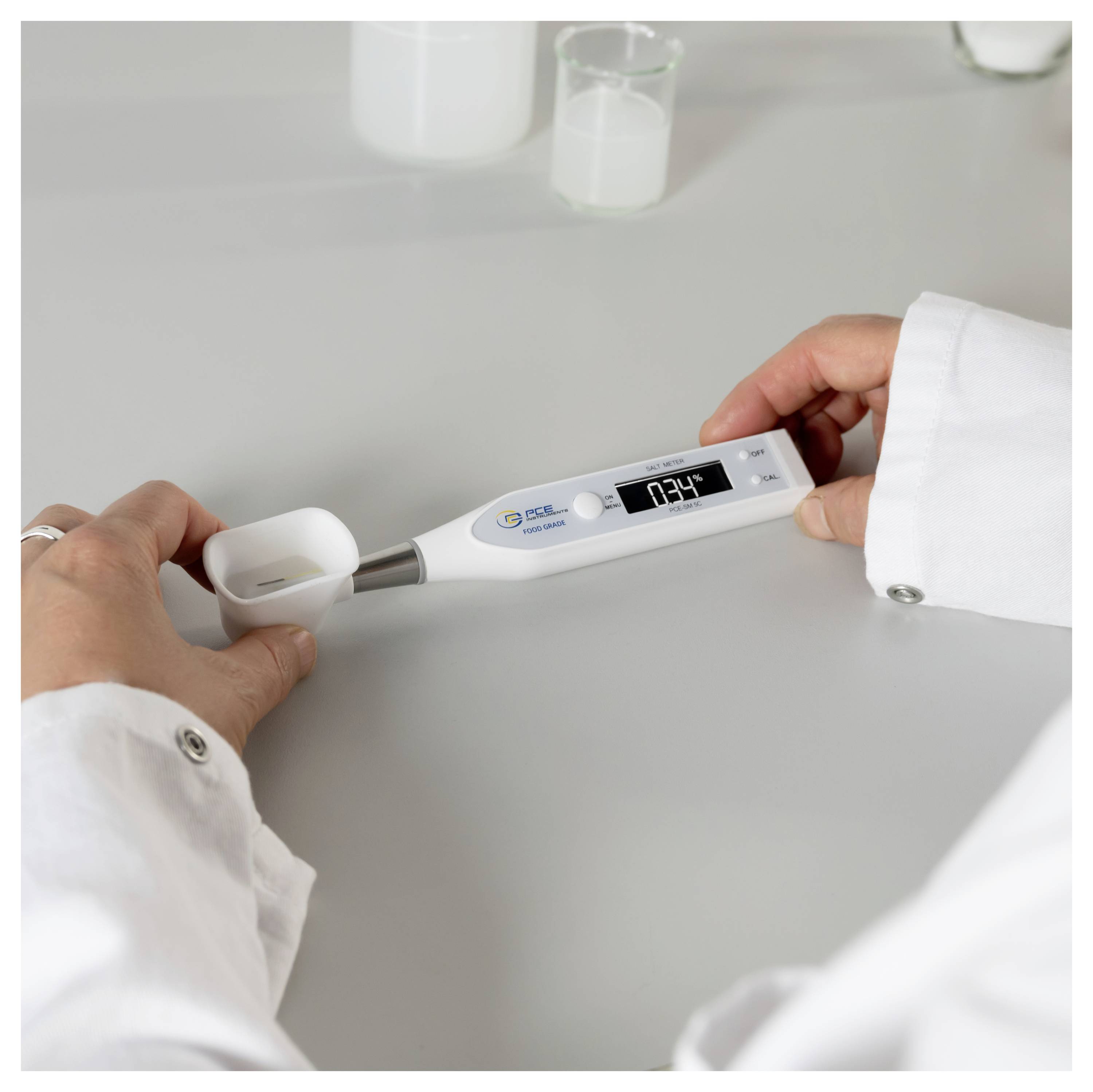 Hände in einem Laborkittel halten ein digitales Thermometer in einer kleinen Tasse auf einer Laborbank, das eine Temperaturanzeige zeigt.