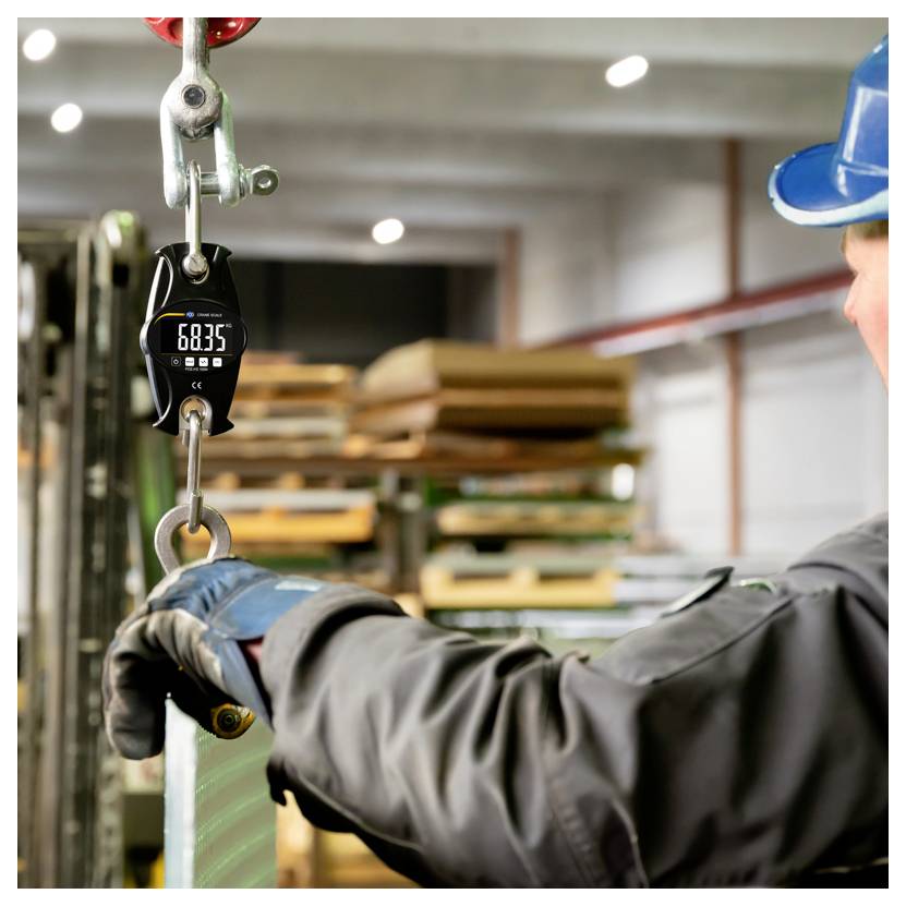 Arbeiter mit Schutzhelm und Handschuhen bedient eine hängende digitale Kranwaage an einer aufgehängten Last in einem Lagerhaus.