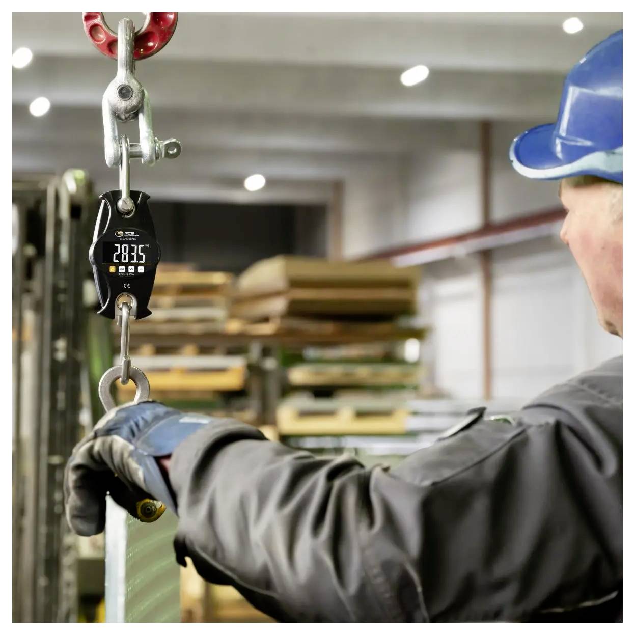 Ein Arbeiter mit Schutzhelm handhabt eine hängende Last mit einer digitalen Kranwaage, die 2835 anzeigt, in einem Industrie-Lagerhaus.