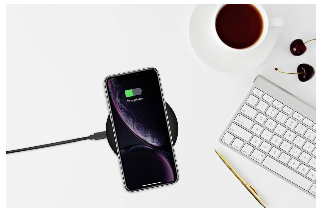 Ein Smartphone, das per Kabel zum Aufladen auf einem weißen Schreibtisch neben einer Tastatur, einem Stift, einer Kaffeetasse und Kirschen angeschlossen ist.