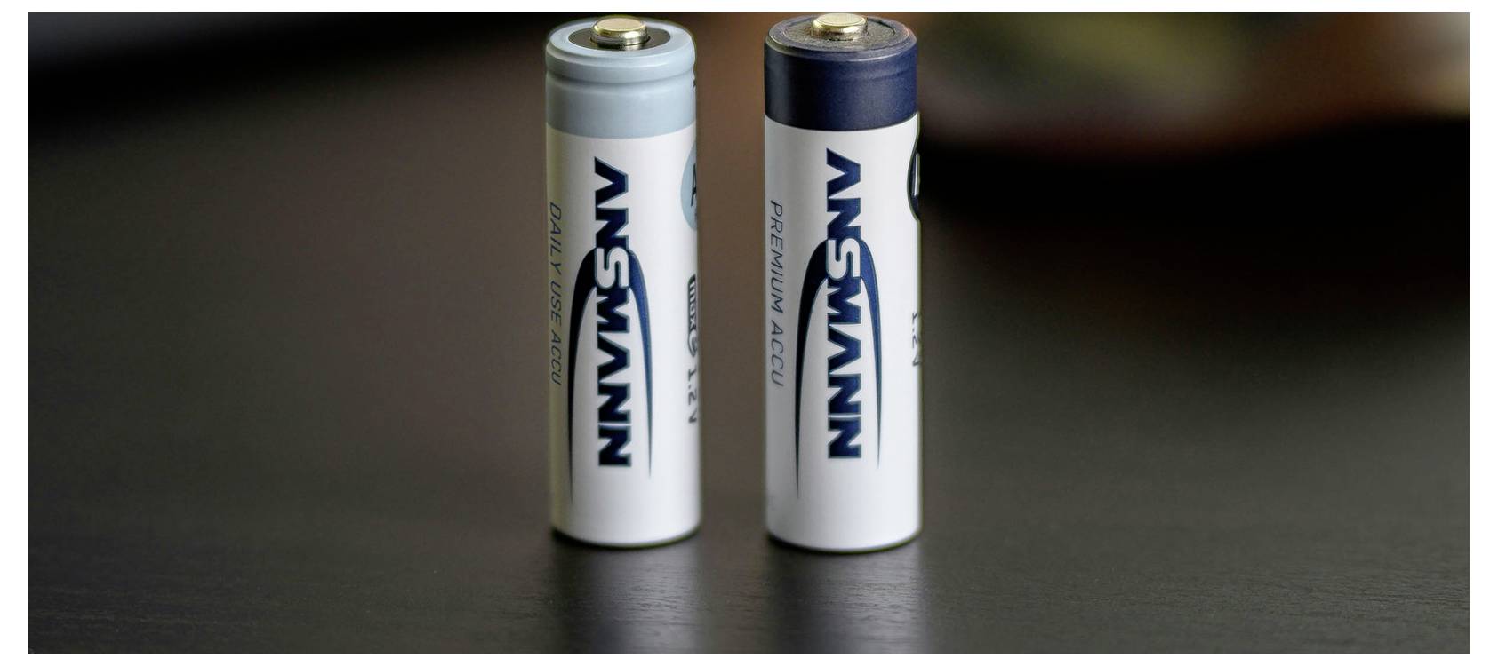 Zwei AA-Batterien, aufrecht auf einer dunklen Oberfläche stehend, eine blau und eine grau, mit dem sichtbaren Markennamen 'ANSMANN'.