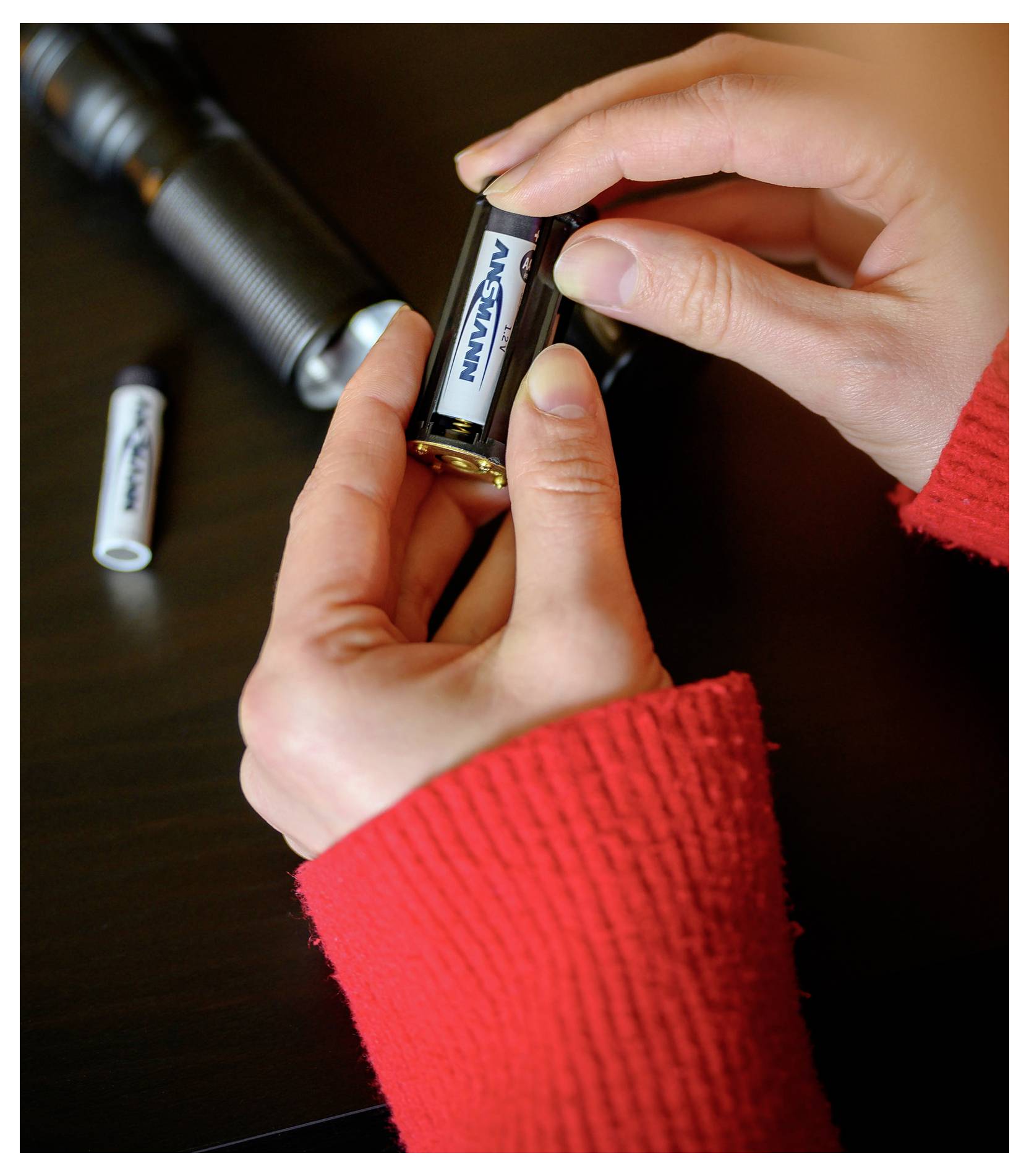 Hände, die die Batterie in einer kleinen Taschenlampe auf einem dunklen Tisch austauschen, mit einer weiteren Batterie in der Nähe.