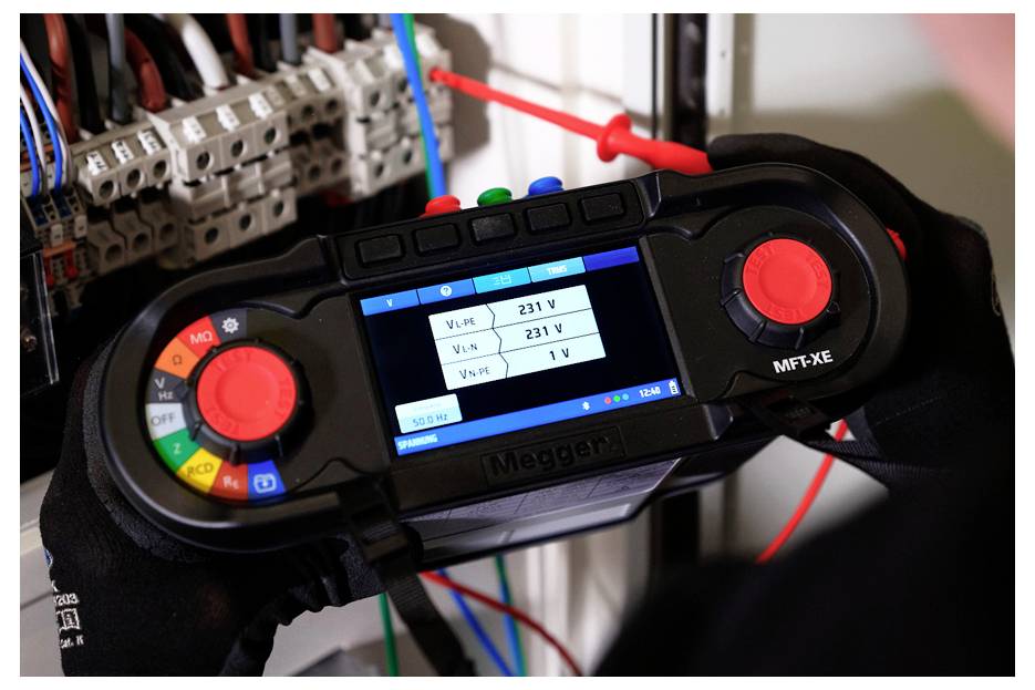 Behandschuhte Hände halten ein Megger-Elektrotester mit Spannungsmessanzeigen in einem Schaltschrank, mit Testleitungen, die an Schutzschaltern angebracht sind.