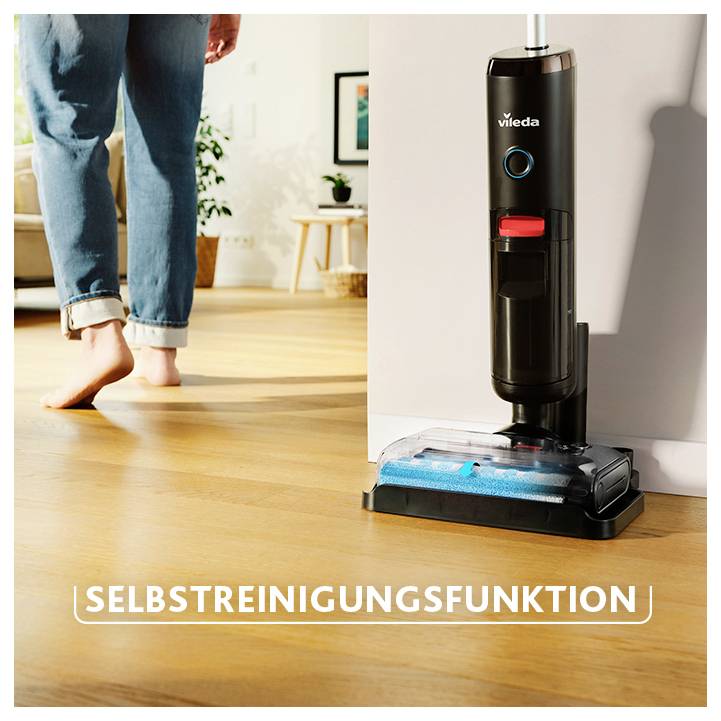 Vileda-Bodenreiniger mit Selbstreinigungsfunktion steht am Holzfußboden; eine Person läuft barfuß vorbei.