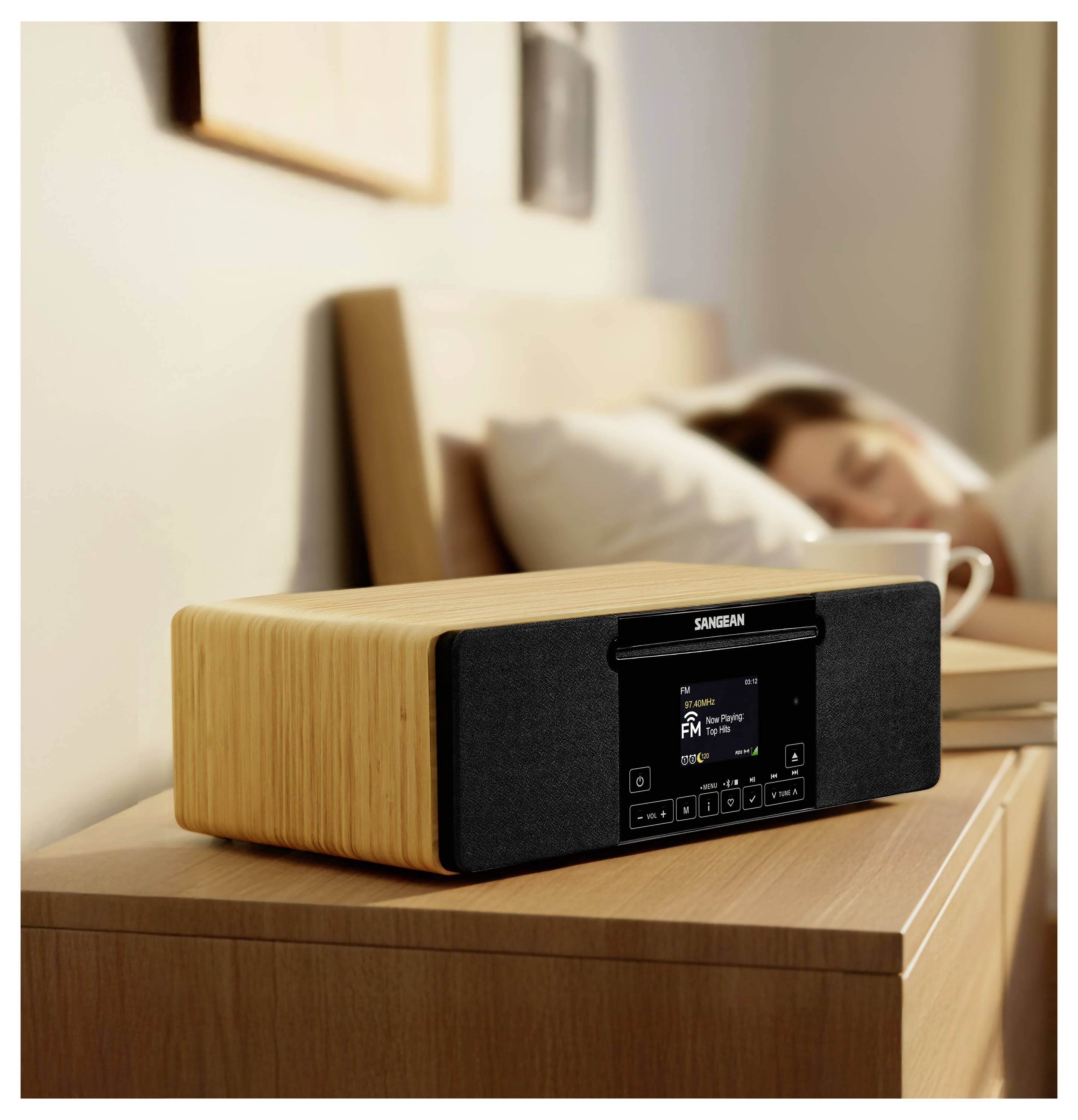 Ein hölzernes digitales Nachttischradio mit Display und Tasten sitzt auf einem Nachttisch, wobei eine schlafende Person verschwommen im Hintergrund zu sehen ist.