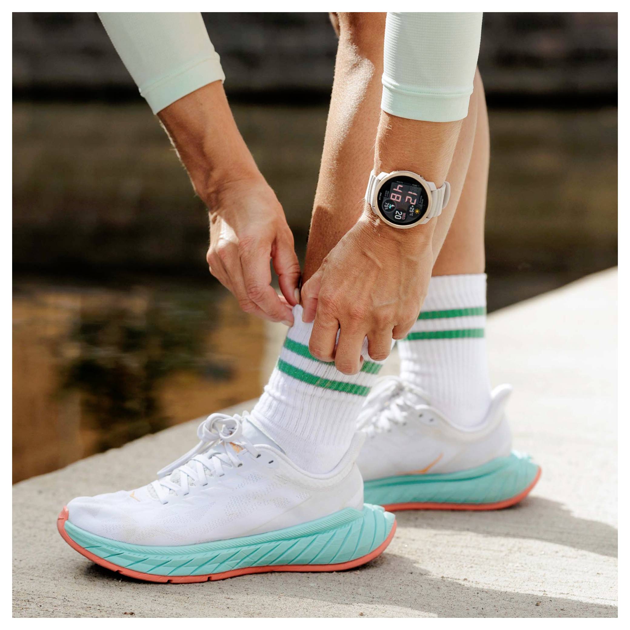 Person, die die Schnürsenkel eines weißen Laufschuhs bindet, trägt gestreifte Socken und eine Smartwatch, draußen neben dem Wasser.