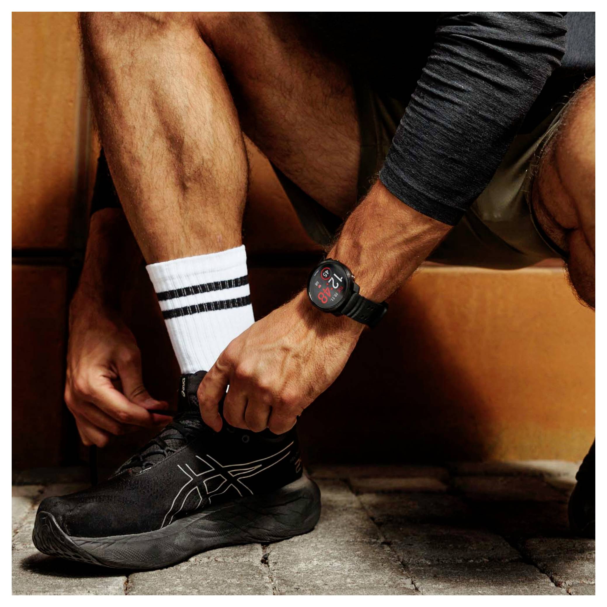 Nahaufnahme eines Mannes, der die Schnürsenkel eines schwarzen Laufschuhs bindet, weiße gestreifte Socken und eine Smartwatch trägt, während er auf einer Bank sitzt.