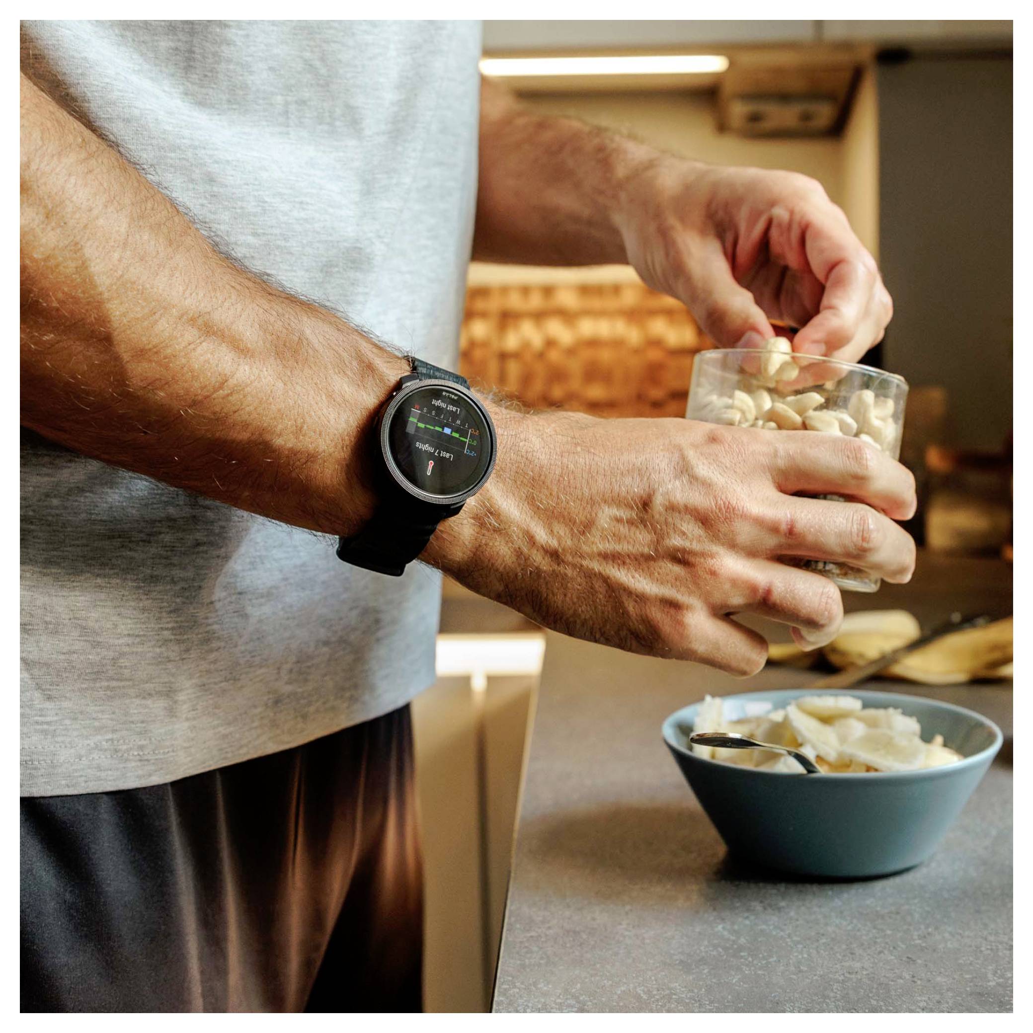 Person, die eine Smartwatch trägt, während sie Nüsse aus einem Behälter in eine blaue Schüssel auf einer Küchentheke gießt.