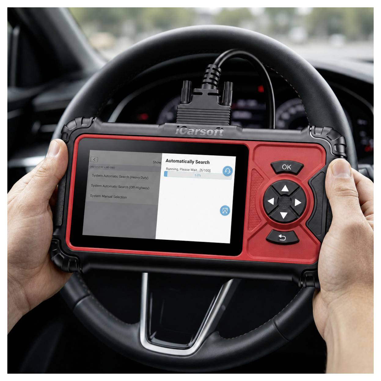 Hände halten ein iCarsoft-Fahrzeugdiagnose-Scan-Gerät vor einem Auto-Lenkrad, das einen "Automatisch Suchen"-Bildschirm anzeigt, während ein System gescannt wird.