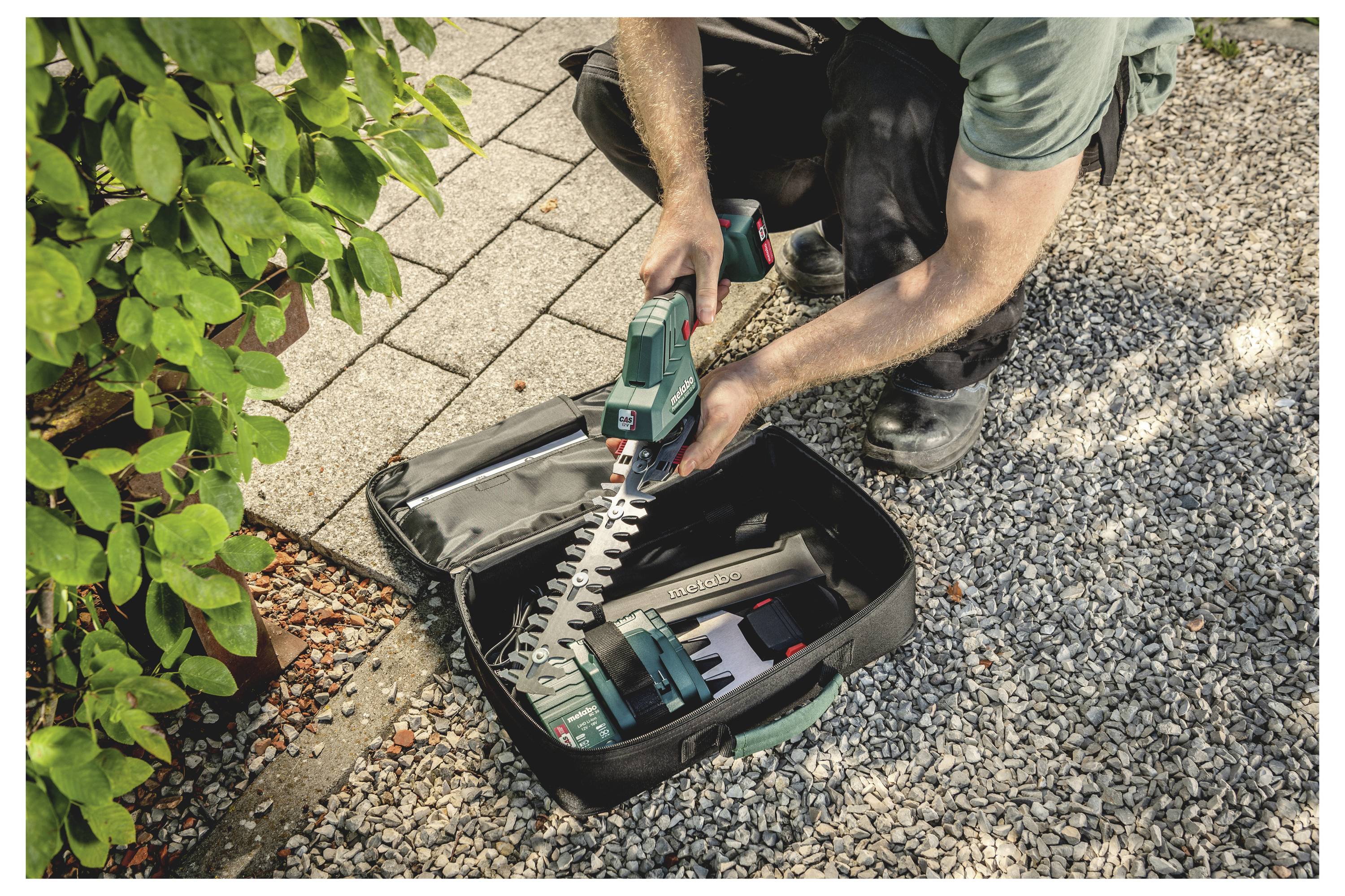 Eine Person trimmt eine Hecke mit einer tragbaren elektrischen Heckenschere, wobei ein offener Werkzeugkoffer auf Kies neben einem gepflasterten Weg liegt.