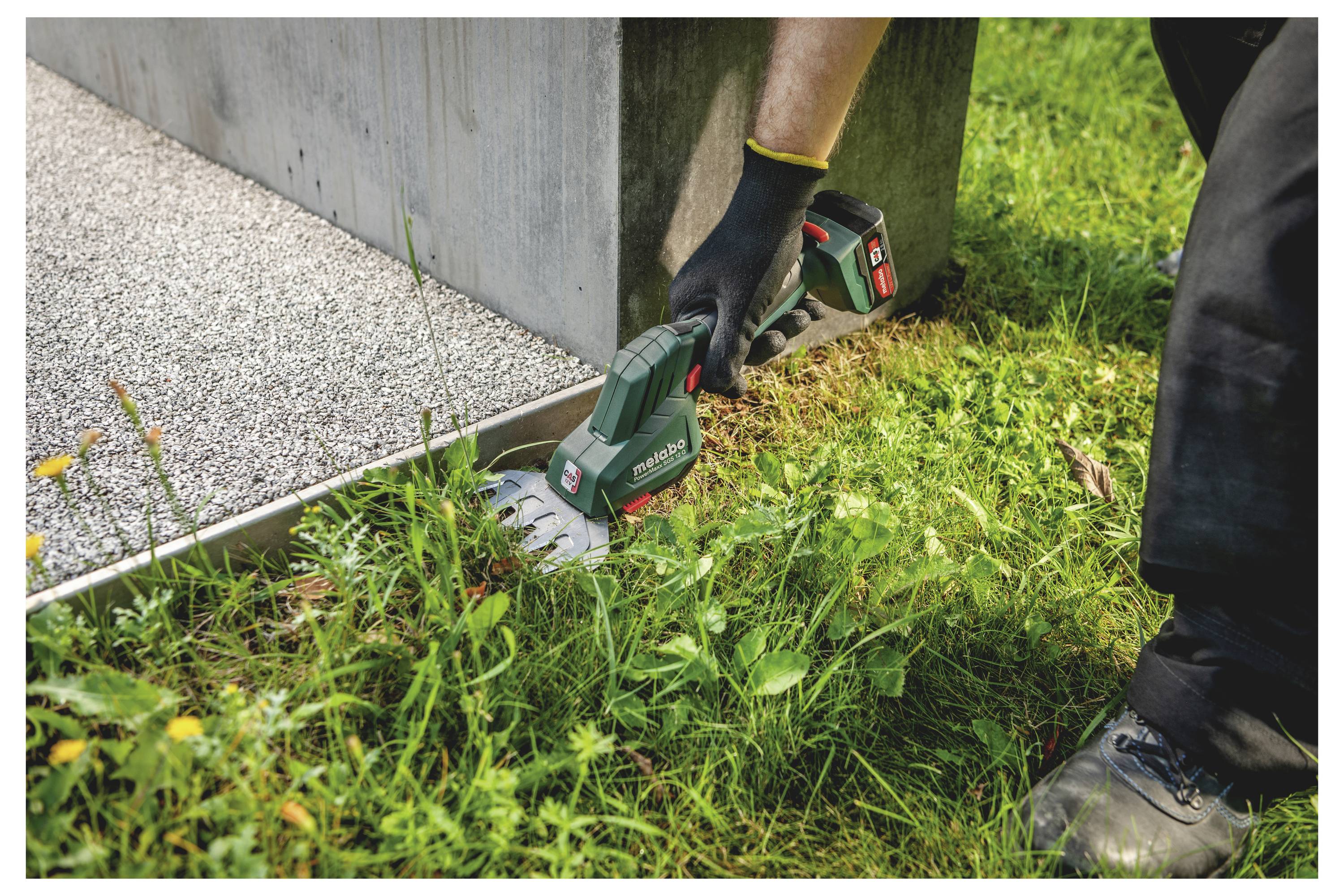 Handschuhgetragener Arbeiter benutzt eine tragbare Elektrosäge, um einen Metallpfosten oder eine Grundplatte neben einer Betonkonstruktion in einem grasbewachsenen Außenbereich zu schneiden.
