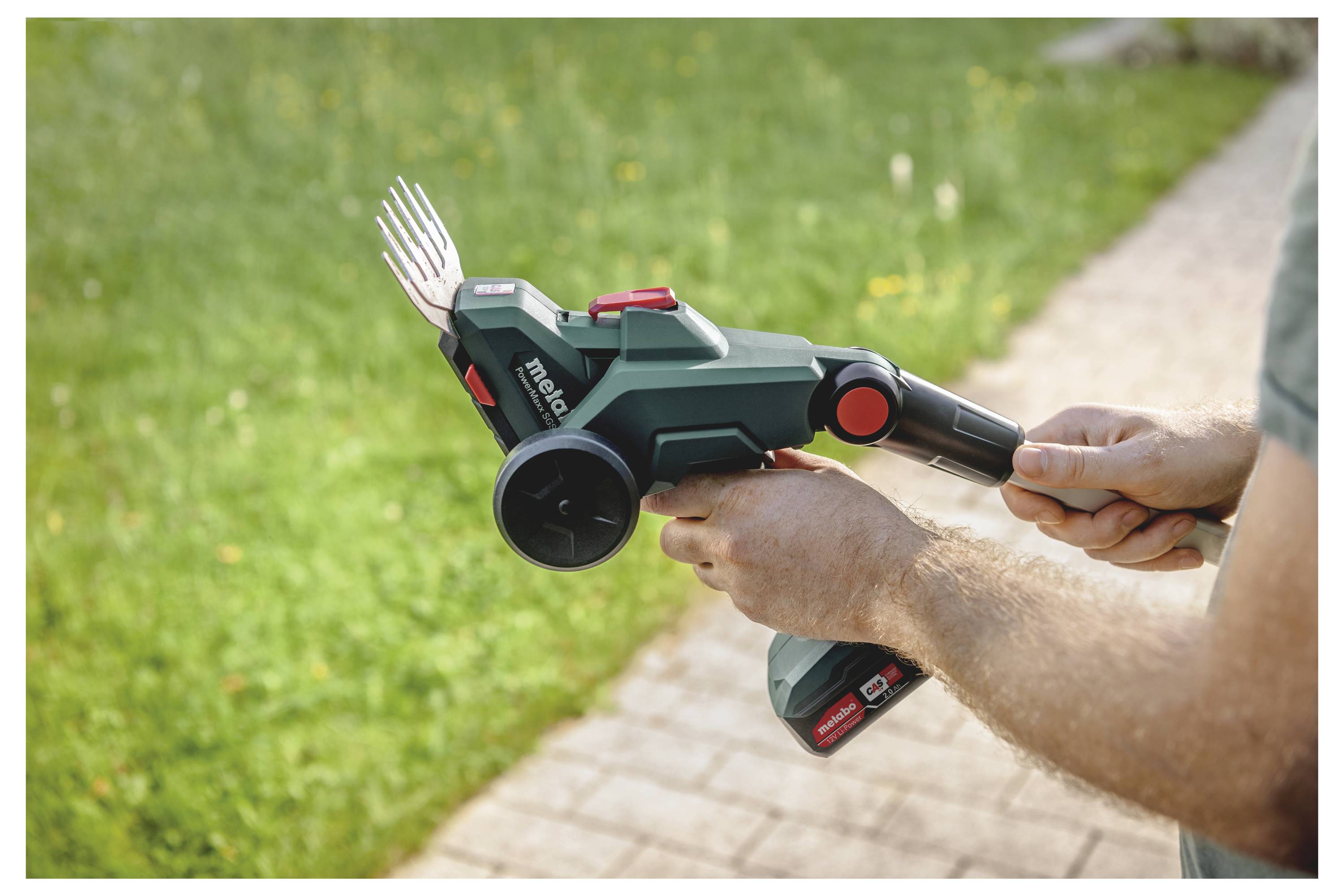 Eine Person benutzt einen schnurlosen Gartenkultivator mit Zinkenaufsatz im Freien auf einem gepflasterten Weg neben Rasen.