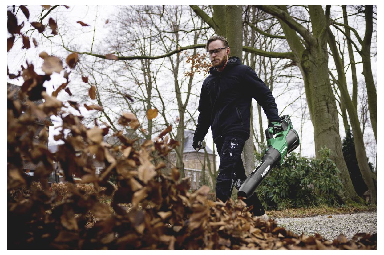 Ein Mann geht im Freien in einem Park und benutzt ein tragbares Laubgebläse, um einen Haufen trockener Herbstblätter von einem Kiesweg zu blasen.