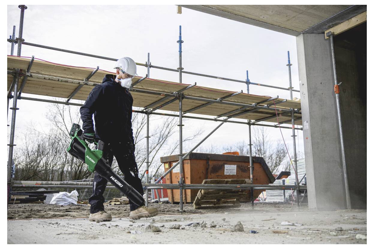 Bauarbeiter mit Schutzhelm und Maske, der eine grüne Elektrosäge verwendet, um Beton auf einer mit Gerüsten beladenen Baustelle zu schneiden.