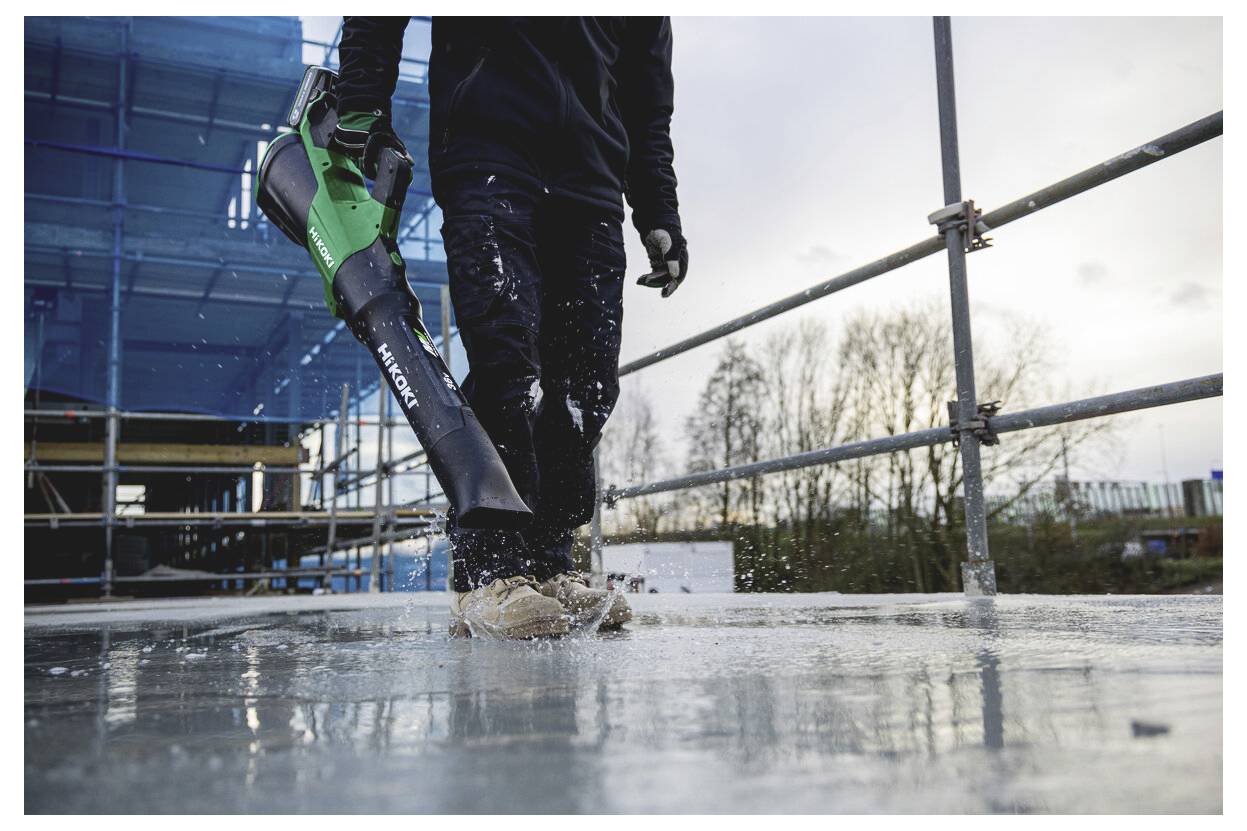 Bauarbeiter geht auf einer nassen Betonplatte, während er ein grün-schwarzes Elektrowerkzeug auf einer Baustelle trägt.