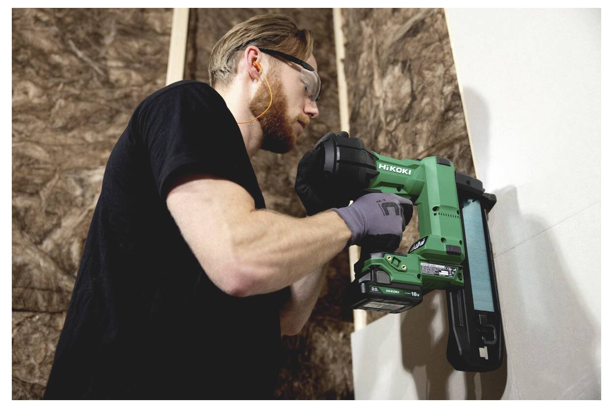Ein Mann benutzt eine grüne kabellose Hitachi-Nagelschussmaschine, um Material an einer Wand während des Innenausbaus zu befestigen, während er eine Schutzbrille und Handschuhe trägt.
