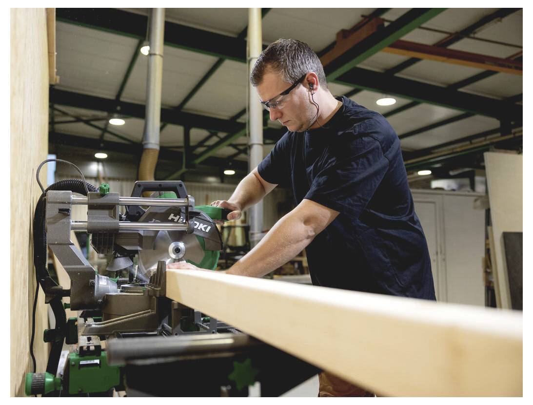 Ein Mann benutzt eine Gehrungssäge, um ein langes Holzbrett in einer Werkstatt zu schneiden, wobei er eine Schutzbrille und Gehörschutzstöpsel trägt.