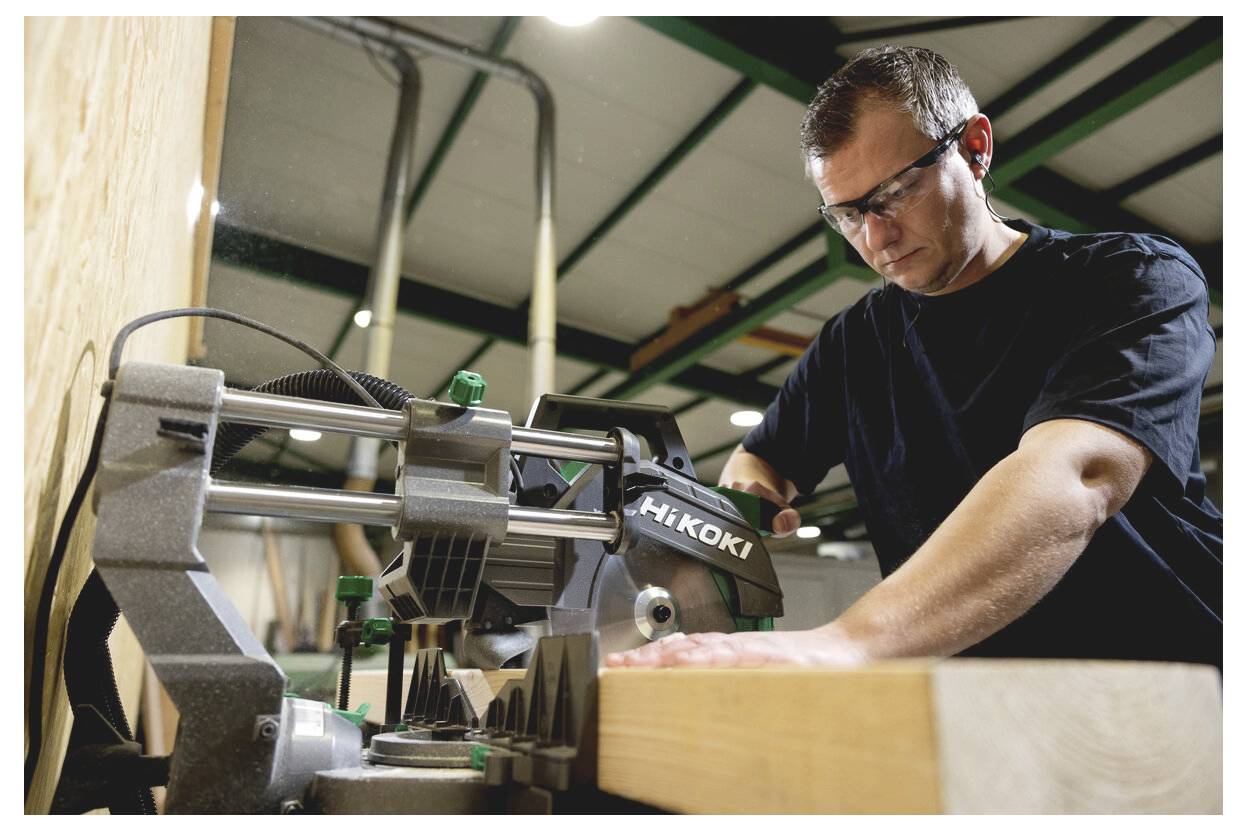 Ein Mann mit einer Schutzbrille bedient eine HIKOKI Elektrosäge in einer Werkstatt und schneidet Holz auf einer Werkbank.