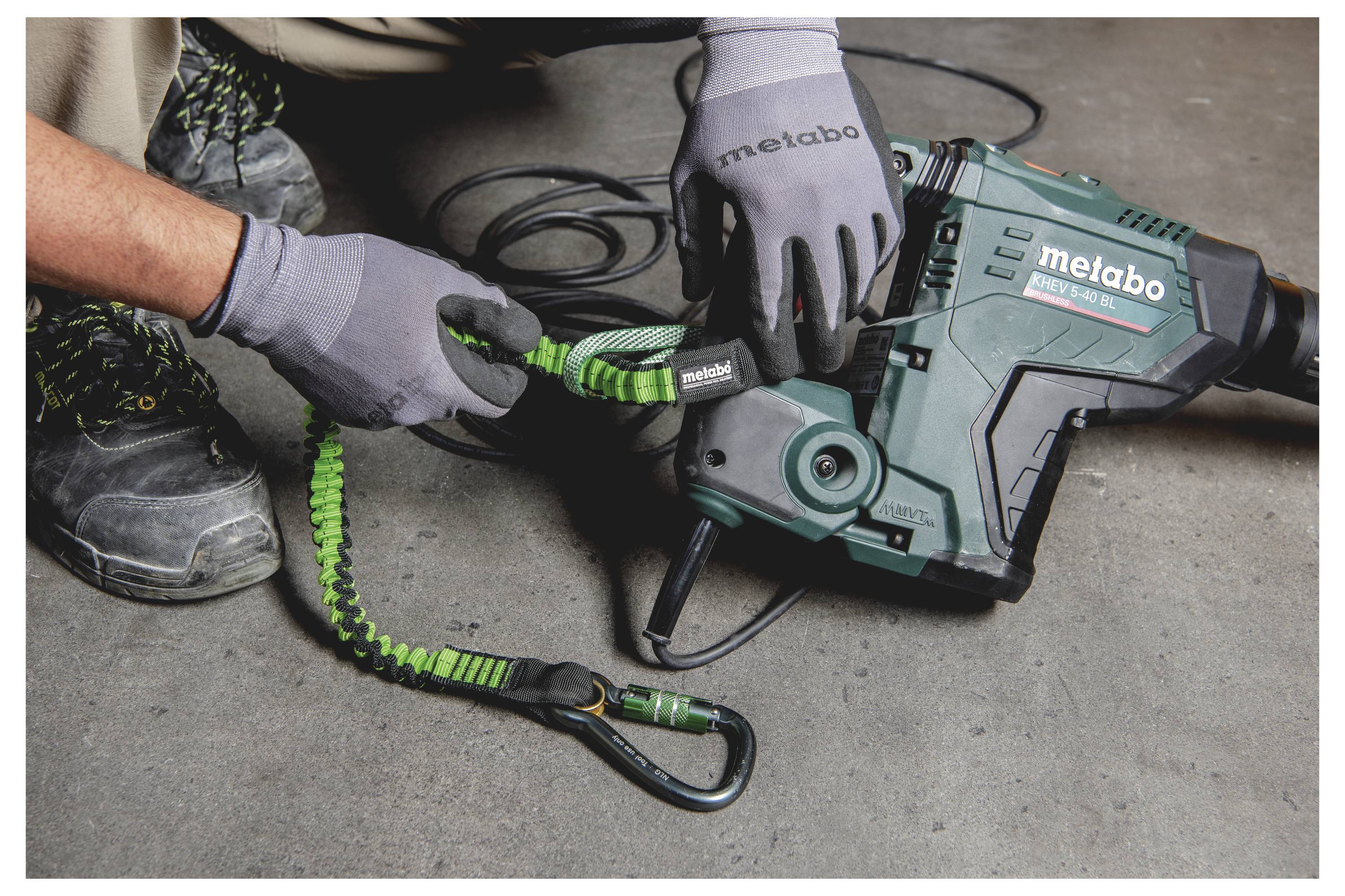 Handschuhgetragener Arbeiter befestigt ein grünes aufgerolltes Verbindungsseil an einem Metabo Elektrowerkzeug auf einem Betonfußboden.
