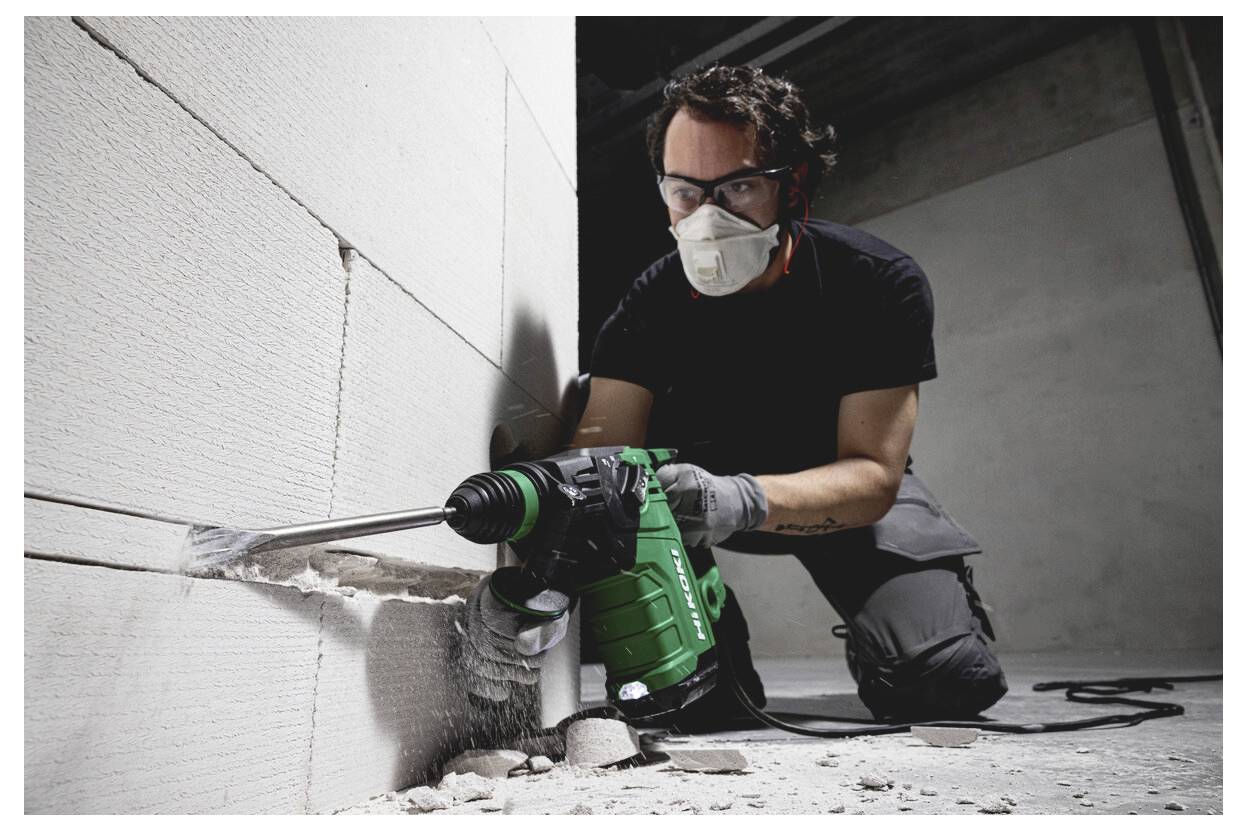Ein Bauarbeiter, der eine Maske und Schutzbrille trägt, bohrt mit einem grünen Elektrowerkzeug in eine Wand und schafft ein Loch in einem unfertigen Innenraum.