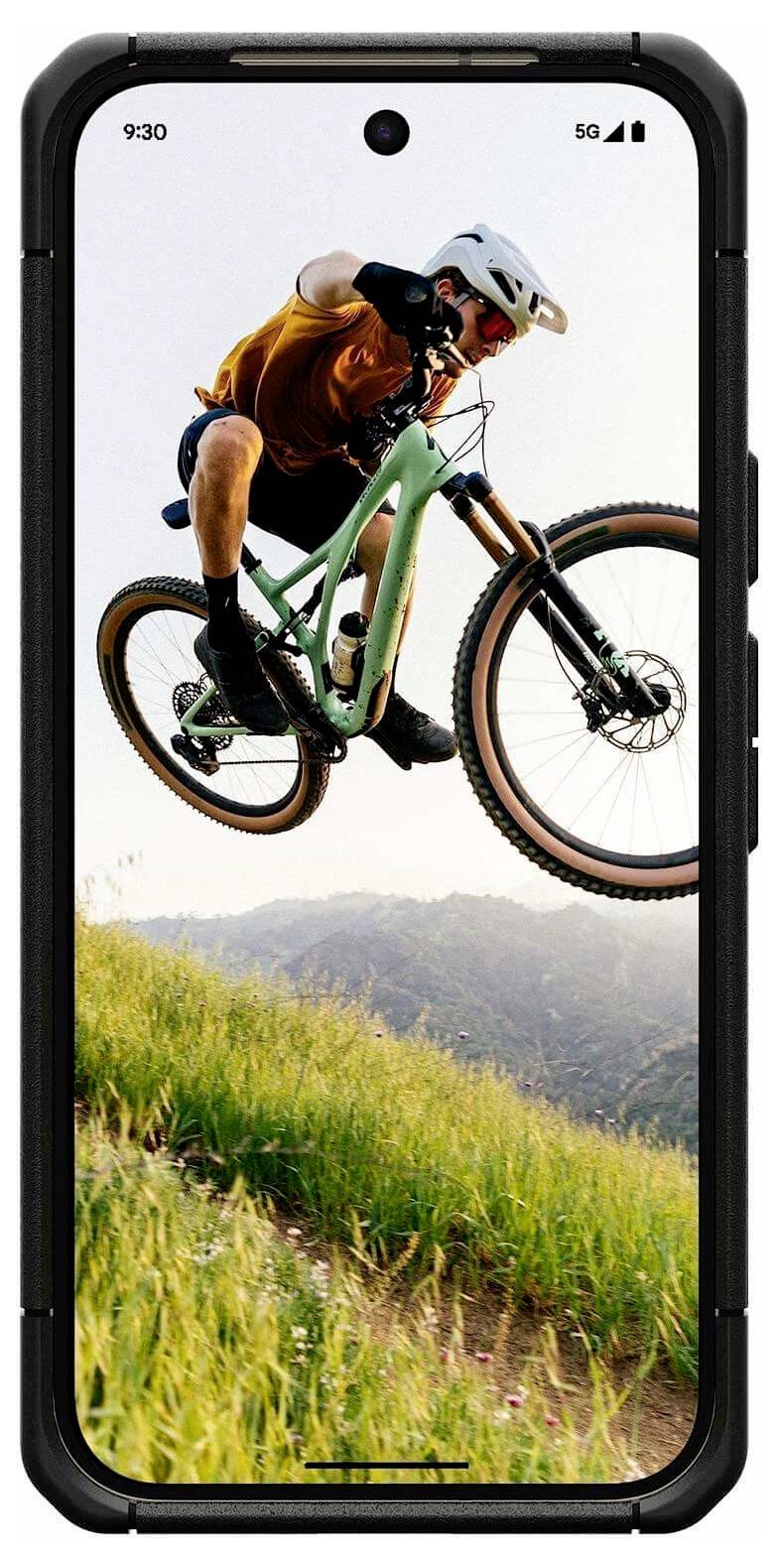 Smartphone-Bildschirm zeigt einen Mountainbiker in der Luft, der einen Sprung über einen grasbewachsenen Hügel macht, mit Hügeln im Hintergrund.
