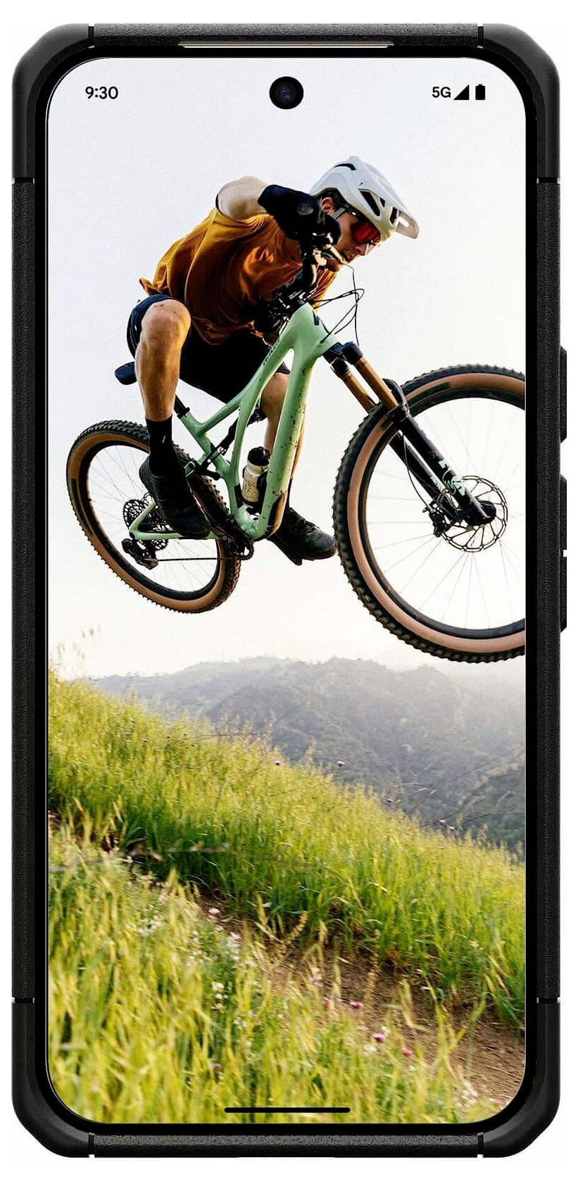 Ein Mountainbiker mitten im Sprung auf einem Trail, schwebend über grasbewachsenen Hügeln bei Sonnenuntergang, angezeigt auf einem Smartphone-Bildschirm.