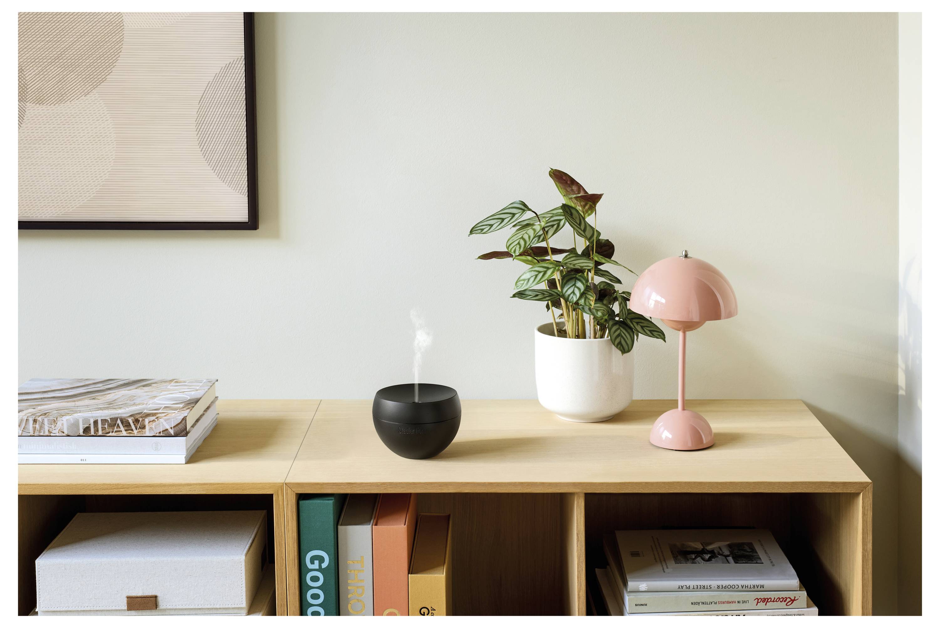 Holzregal mit Büchern, Aufbewahrungsboxen, einer Topfpflanze, einer rosafarbenen Tischlampe und einem kleinen schwarzen Luftbefeuchter oder Diffusor, der Dampf gegen eine helle Wand abgibt.