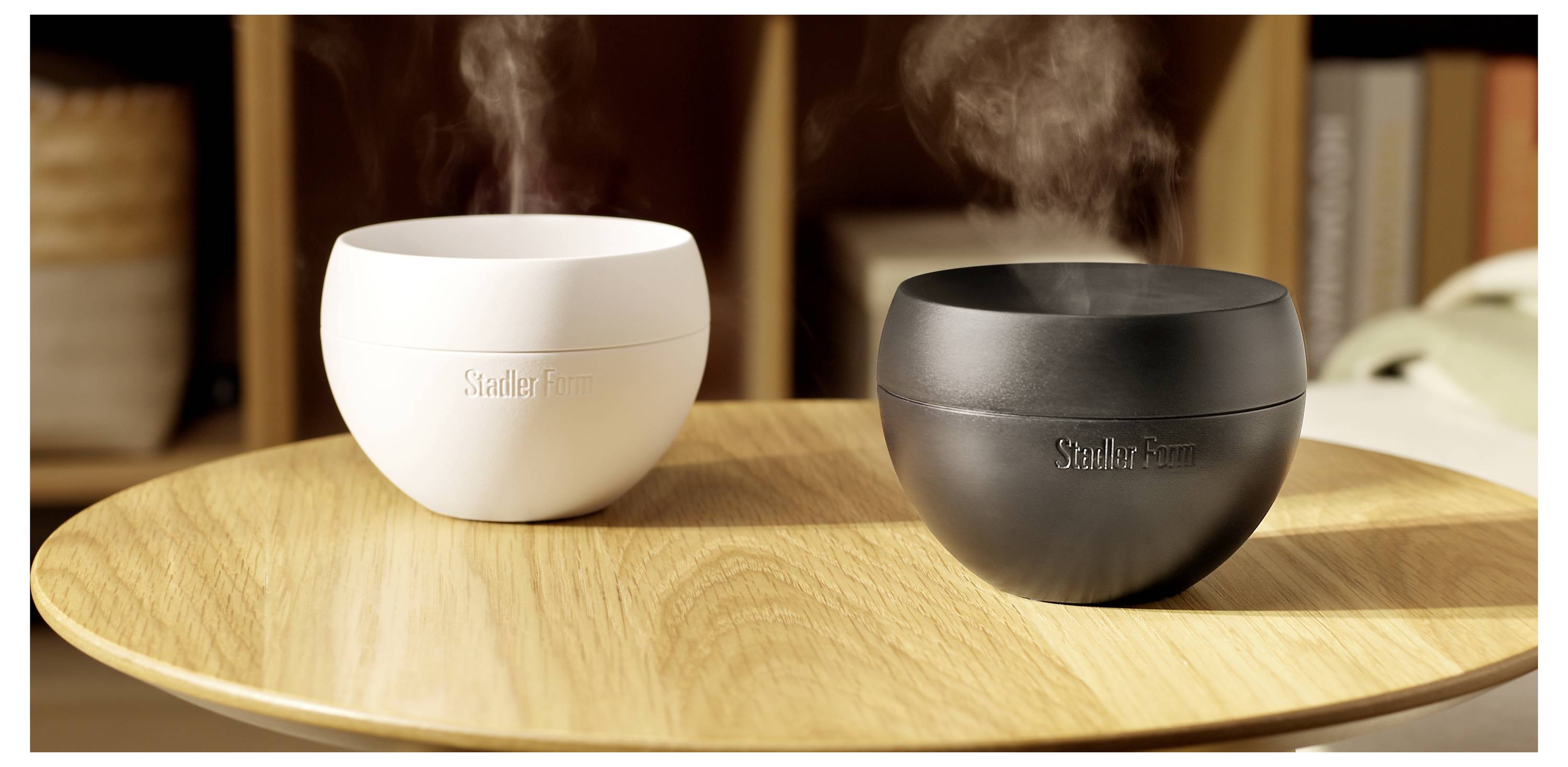 Zwei runde Tischplatten-Aromadiffusoren, einer weiß und einer schwarz, geben sichtbaren Dampf auf einem Holztablett ab.