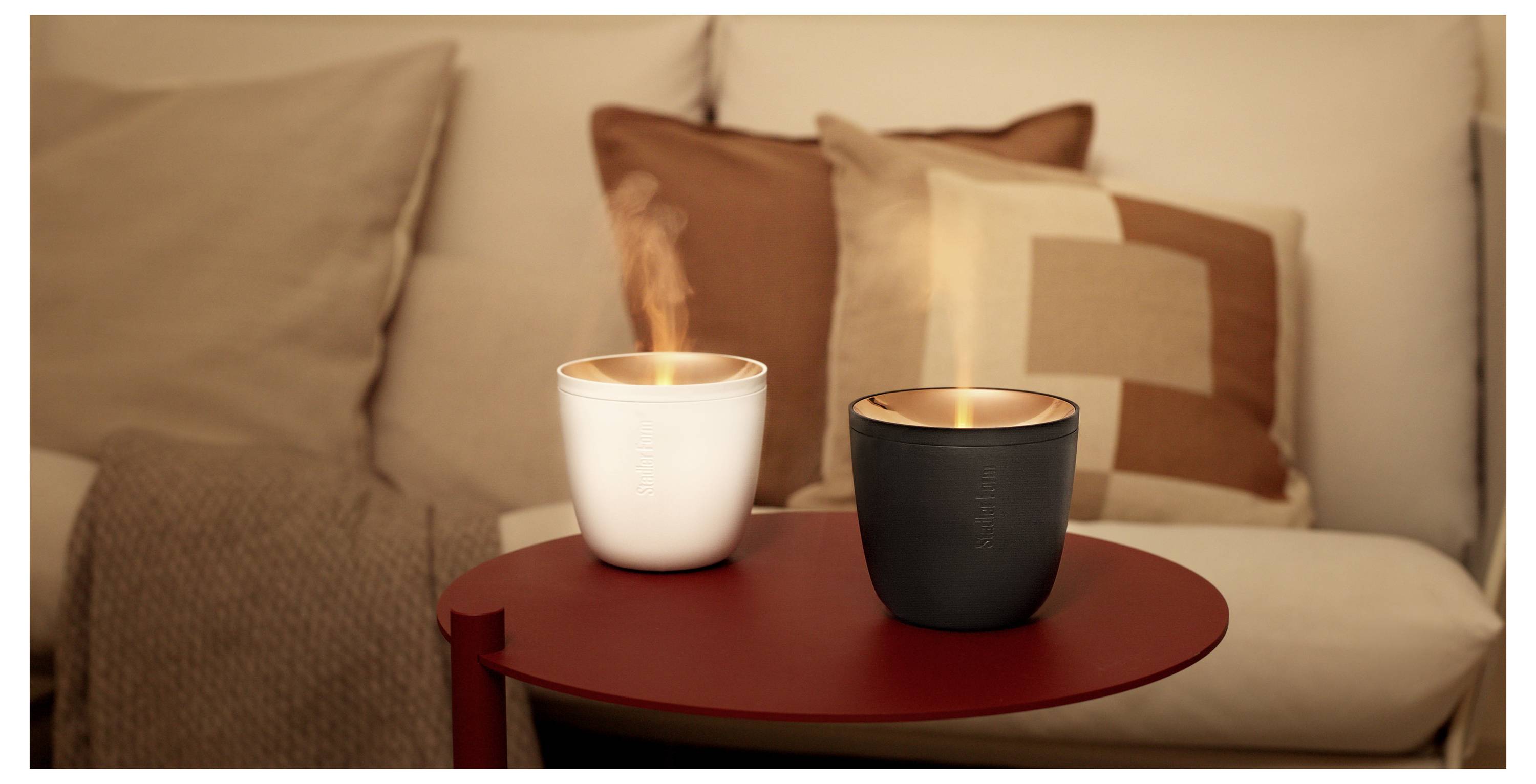 Zwei brennende Kerzen in weißen und schwarzen Kerzenhaltern auf einem roten Tisch, mit gemütlichen Sofakissen im Hintergrund.