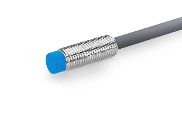 Nahaufnahme eines zylindrischen Näherungssensors mit blauer Spitze und gewindetem Metallgehäuse, das an einem grauen Kabel an einem weißen Hintergrund befestigt ist.