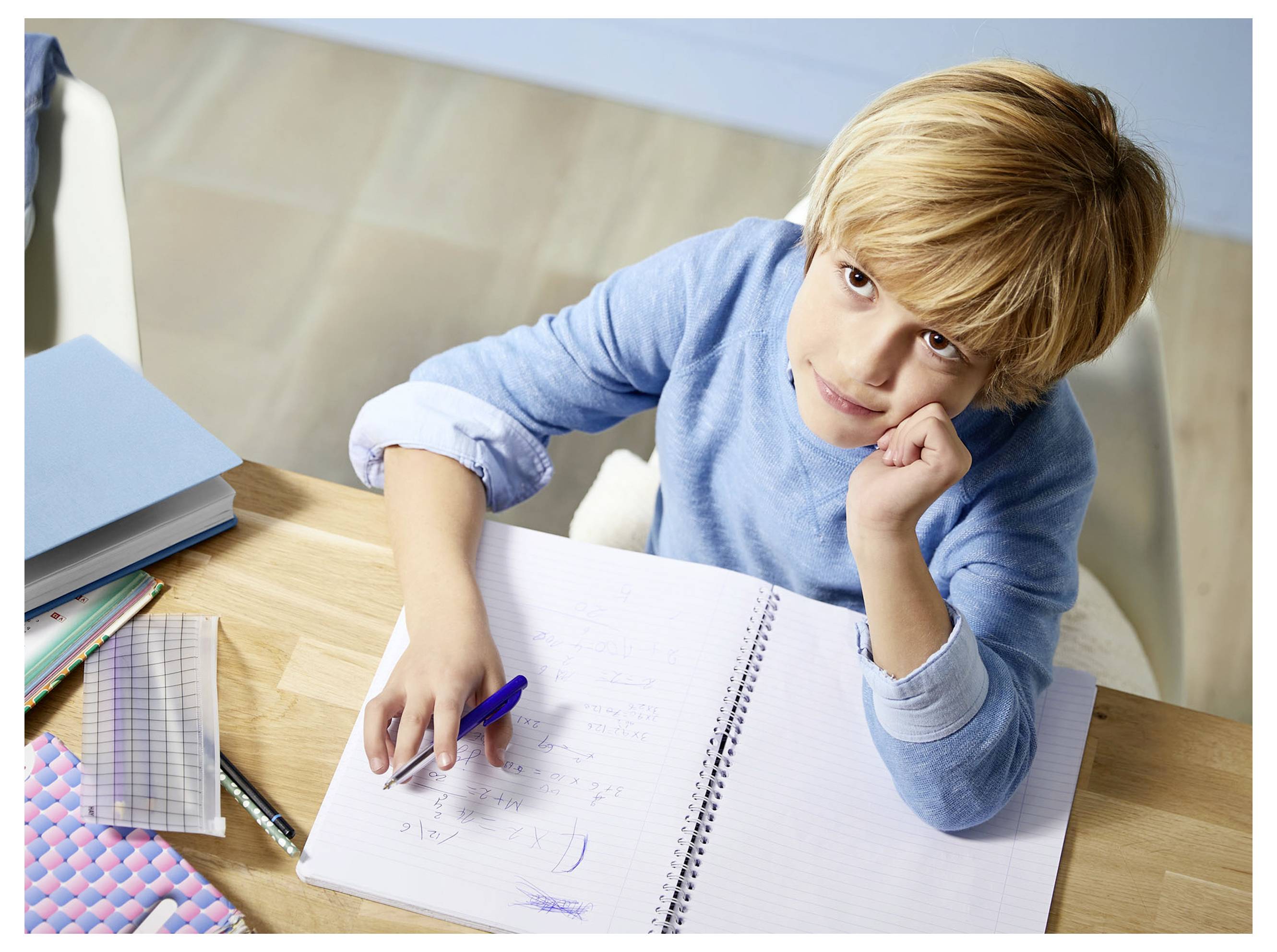 Ein junger Junge sitzt an einem Schreibtisch mit einem offenen Notizbuch, hält einen Stift und denkt nach, während er lernt oder seine Hausaufgaben macht.