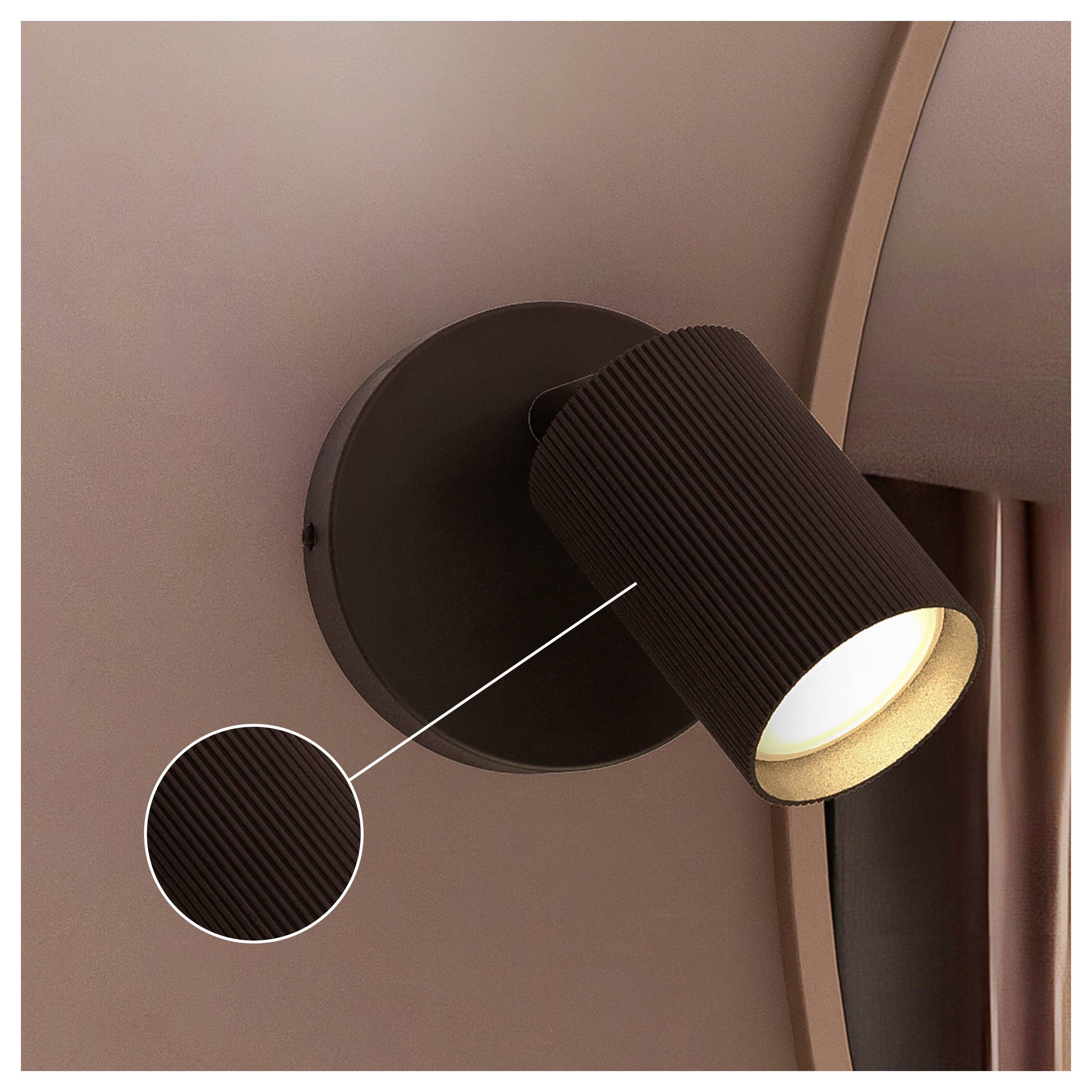 Schwarze zylindrische Wandleuchte mit geripptem Schirm, beleuchtet und nach unten geneigt an einer braunen Wand.