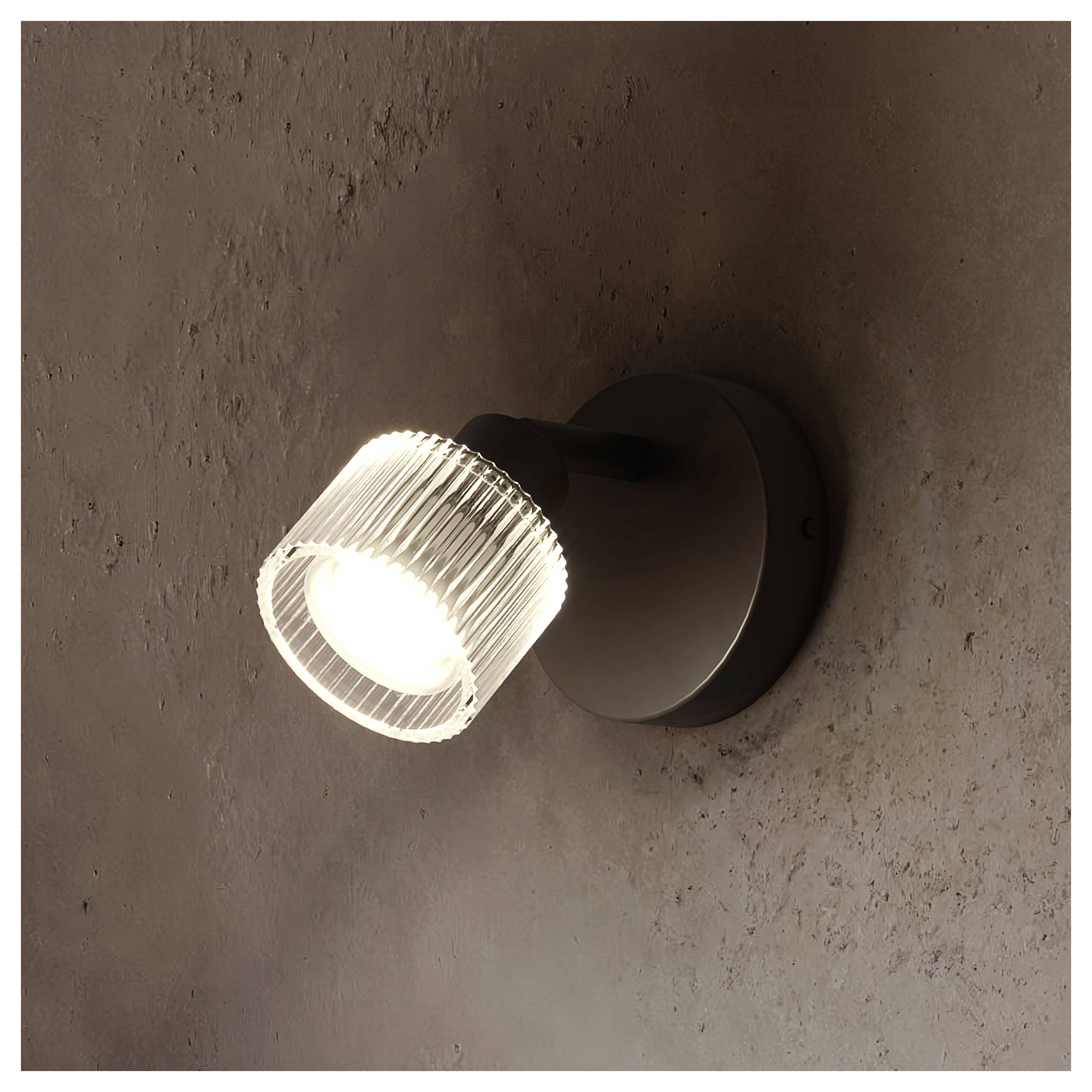 Wandmontierter Scheinwerfer mit einer leuchtenden gerippten Glasleuchte auf einer dunklen kreisförmigen Basis vor einer strukturierten braunen Wand.