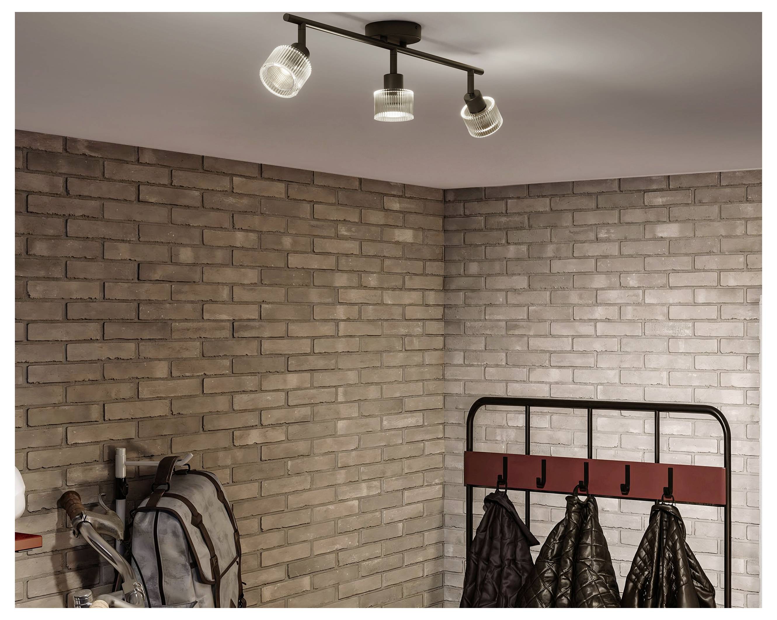 Leere Ecke mit beigen Ziegelmauern, Deckenspot, Rucksack und Garderobe mit Jacken.