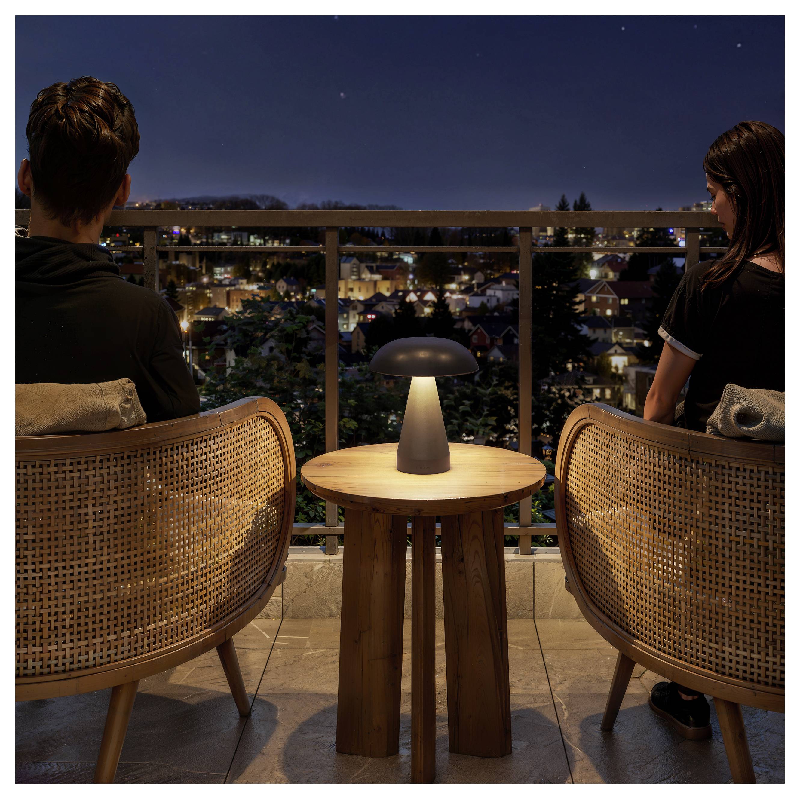Zwei Menschen sitzen nachts auf einem Balkon und blicken auf ein beleuchtetes Wohnviertel einer Stadt. Auf dem Tisch zwischen ihnen brennt eine kleine Lampe.