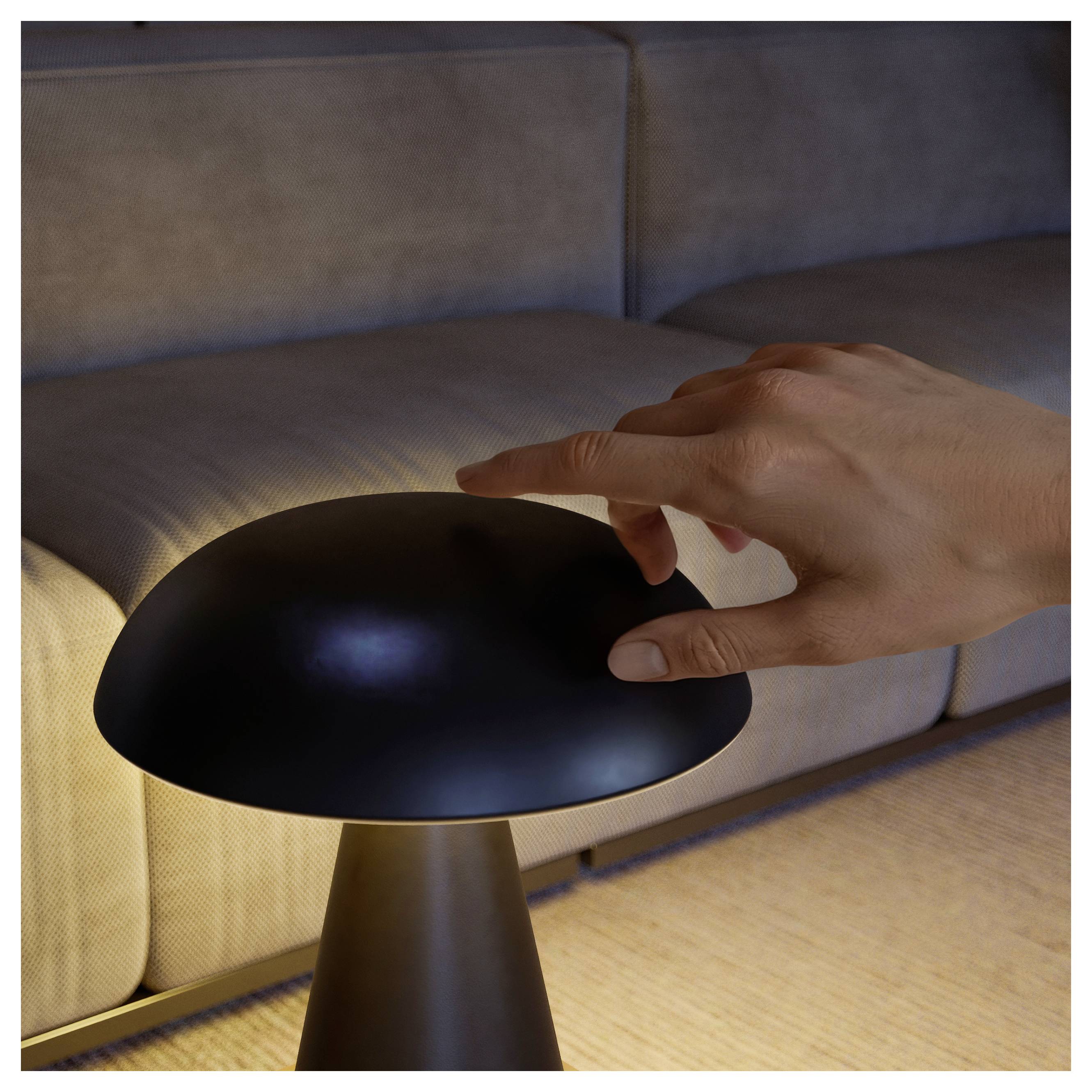 Hand over einer kuppelförmigen schwarzen Lampe auf einem Tisch neben einem Sofa in einem sanft beleuchteten Raum schwebend.