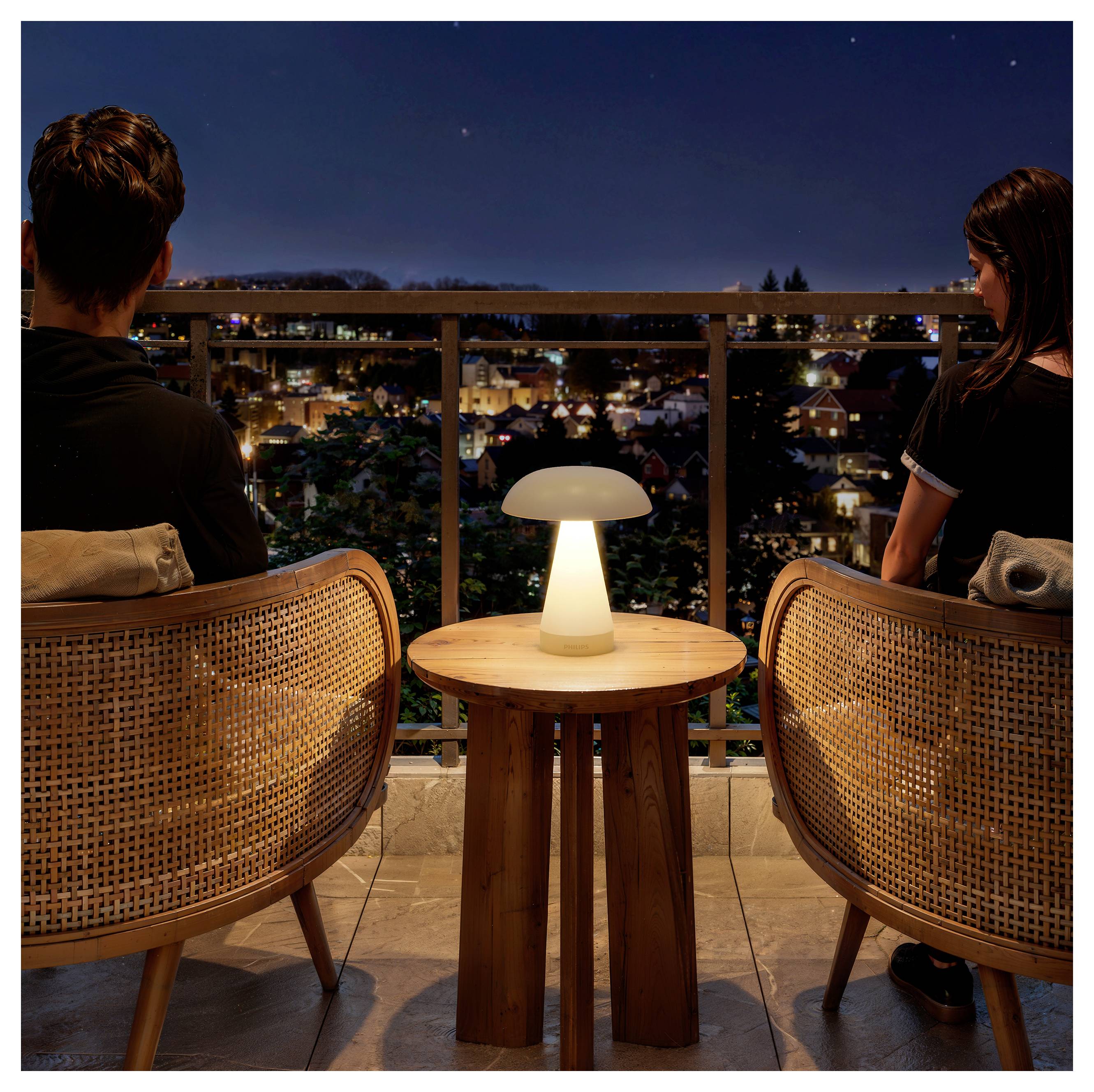 Zwei Menschen sitzen nachts auf einem Balkon und blicken auf eine beleuchtete Stadtlandschaft, während zwischen ihren Stühlen eine kleine leuchtende Tischlampe steht.