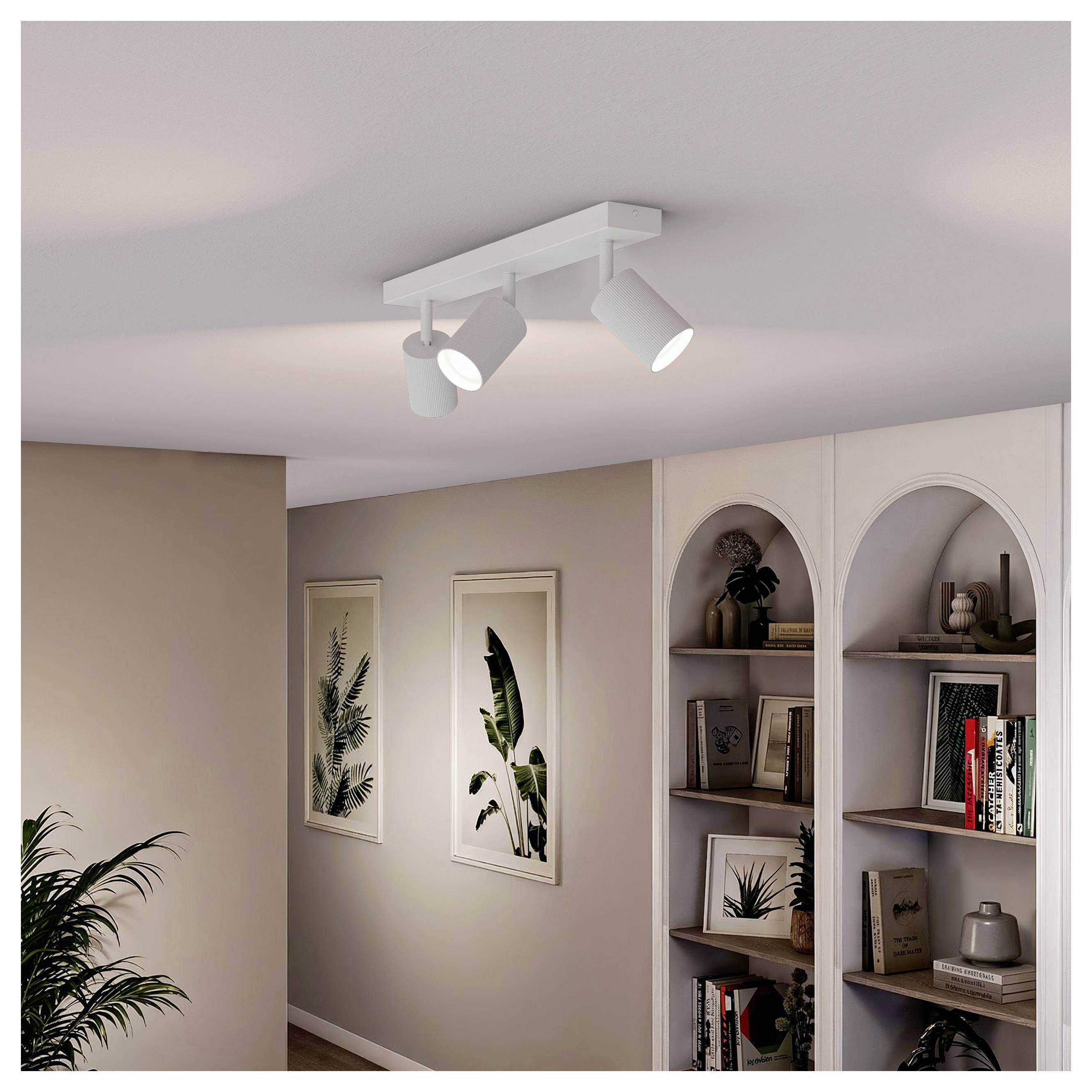 Weiße Deckenleuchte mit Schiene mit drei verstellbaren Spotlights in einem beigen Zimmer, neben gewölbten Wandregalen und gerahmten Pflanzenmotiven.