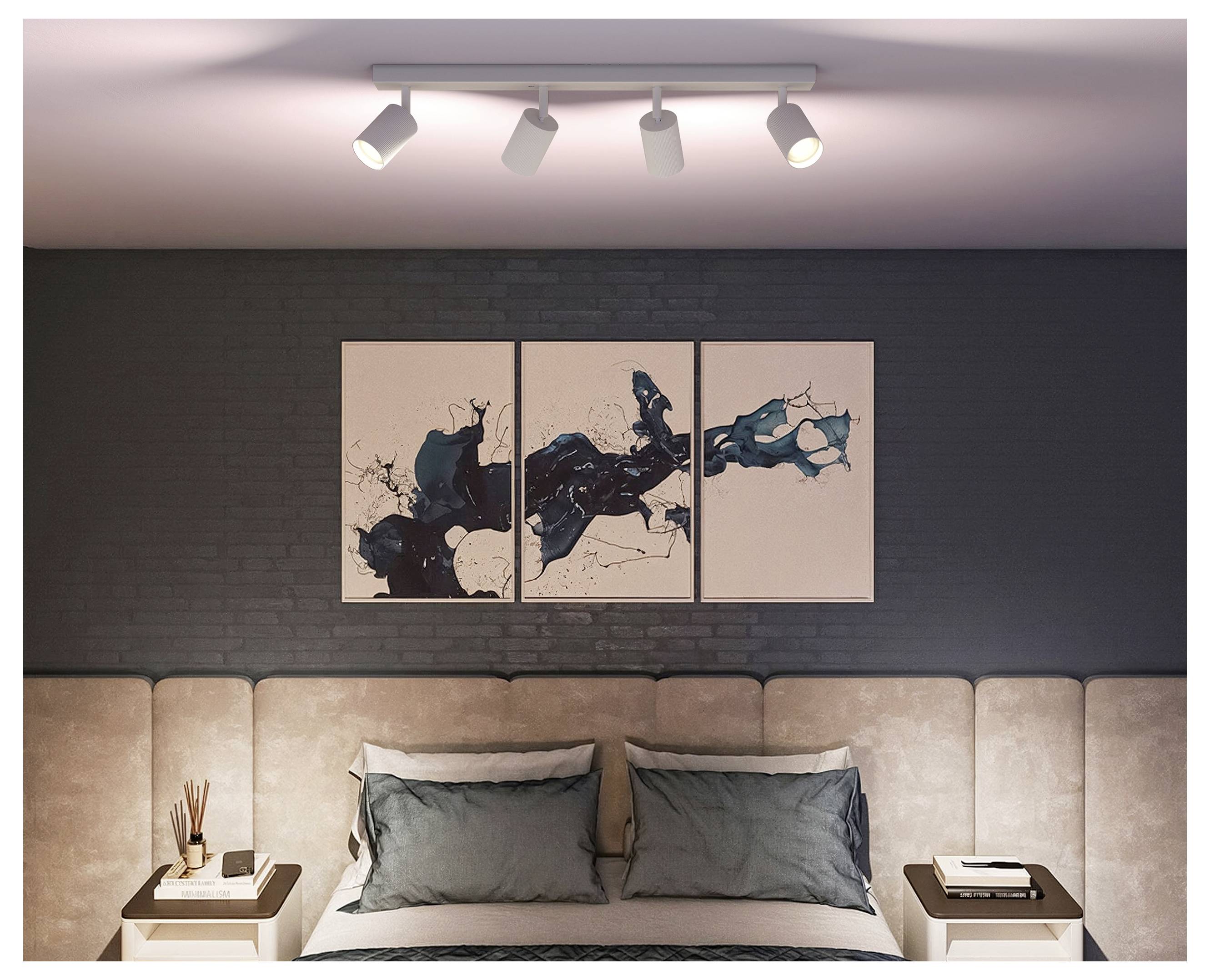 Modernes Schlafzimmer mit einem beigen gepolsterten Bett, grauen Kissen, Nachttischen, Schienenleuchten und abstraktem dreiteiligem Kunstwerk an einer dunklen Backsteinwand.