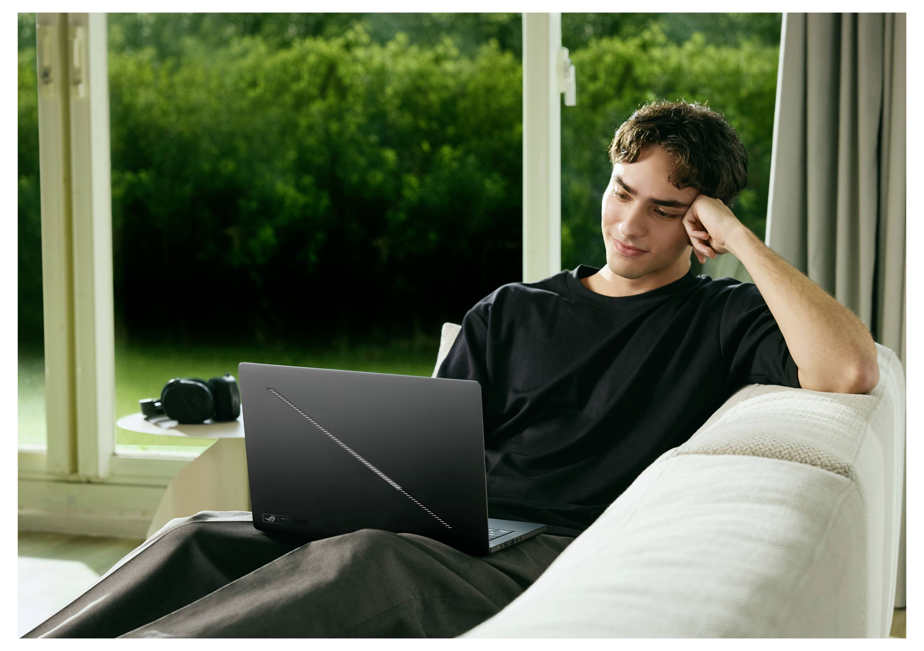 Junger Mann, der sich auf einem Sofa entspannt, mit einem Laptop auf seinem Schoß, schaut auf den Bildschirm in einem hellen Raum neben großen Fenstern.