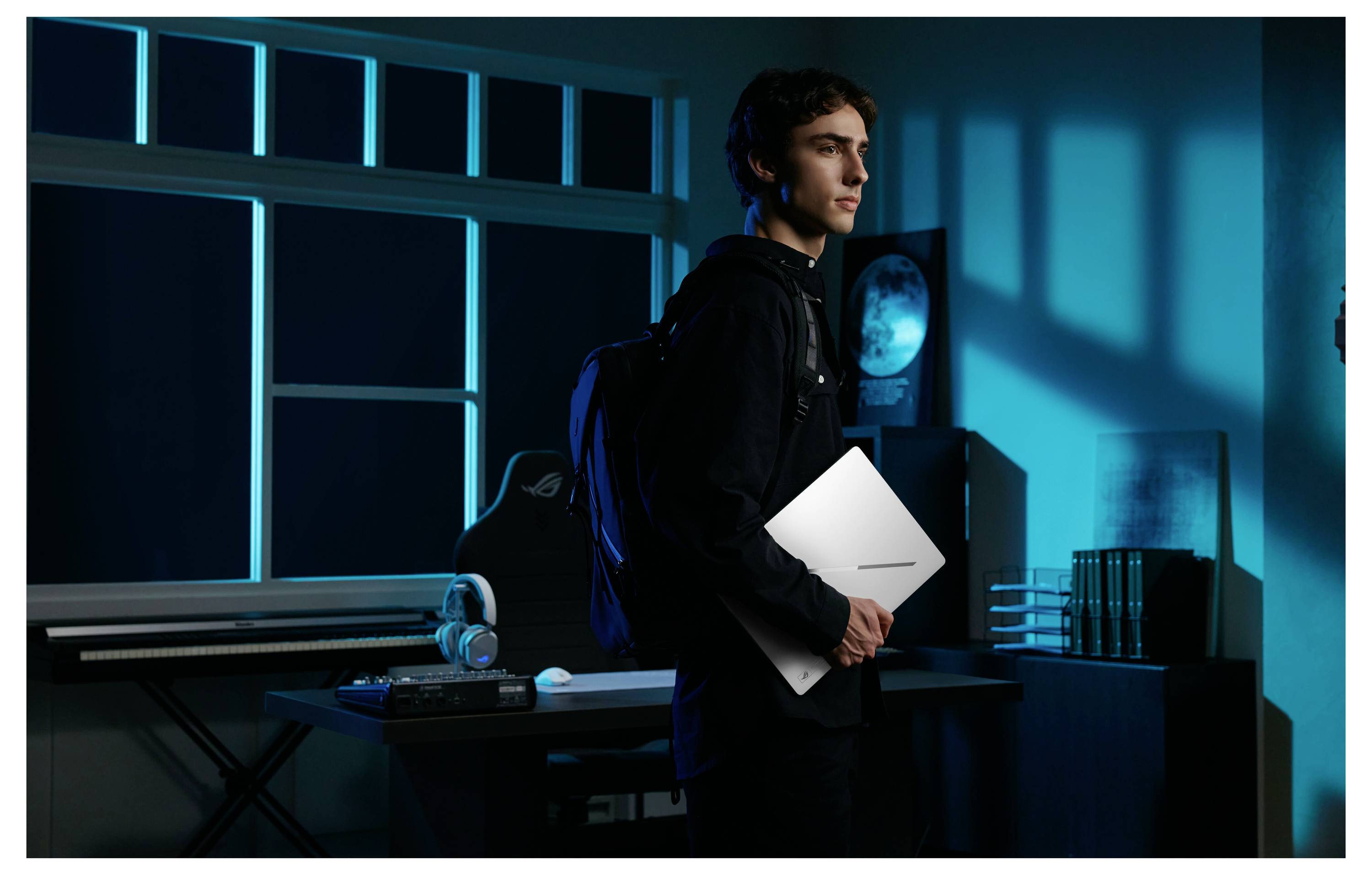 Junger Mann, der einen weißen Laptop in einem blau beleuchteten Gaming- oder Computerraum hält, der neben einem Schreibtisch mit Kopfhörer und Monitor steht.