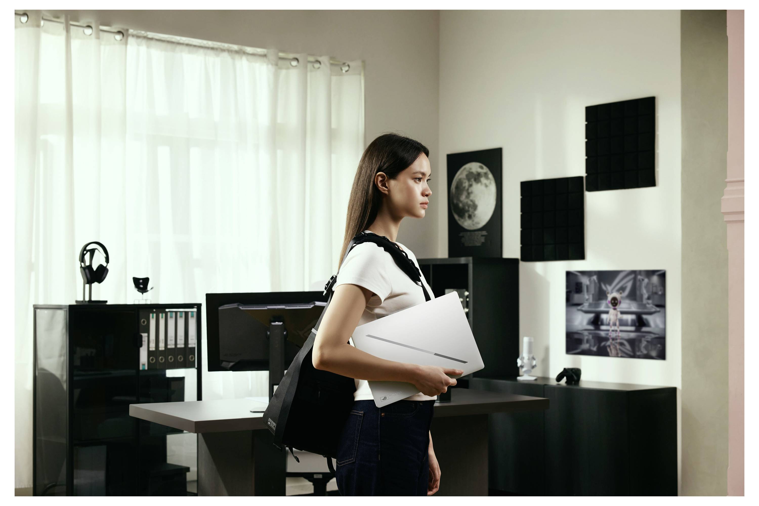 Frau mit Rucksack und Ordner, die in einem modernen Büro oder Studio mit Schreibtischen, Monitoren, Regalen und Wandkunst steht.
