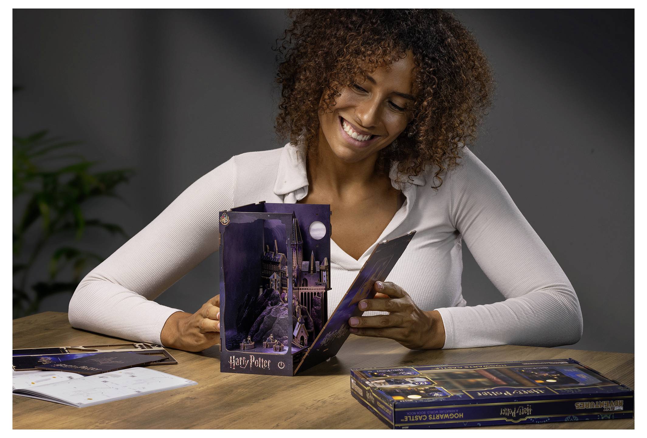 Lächelnde Frau untersucht eine Harry-Potter-Brettspielbox auf einem Tisch, mit Spielfiguren und einer Anleitung, die vor ihr ausgebreitet sind.