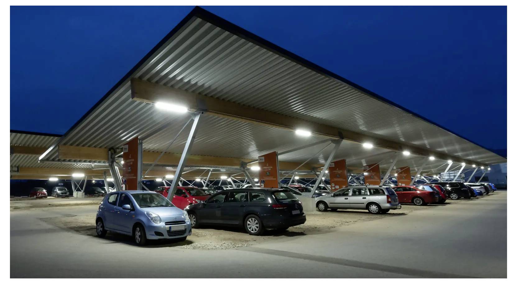 Nächtliche Ansicht eines modernen überdachten Parkplatzes mit mehreren geparkten Autos unter einem großen Metallvordach, das von Deckenleuchten beleuchtet wird.