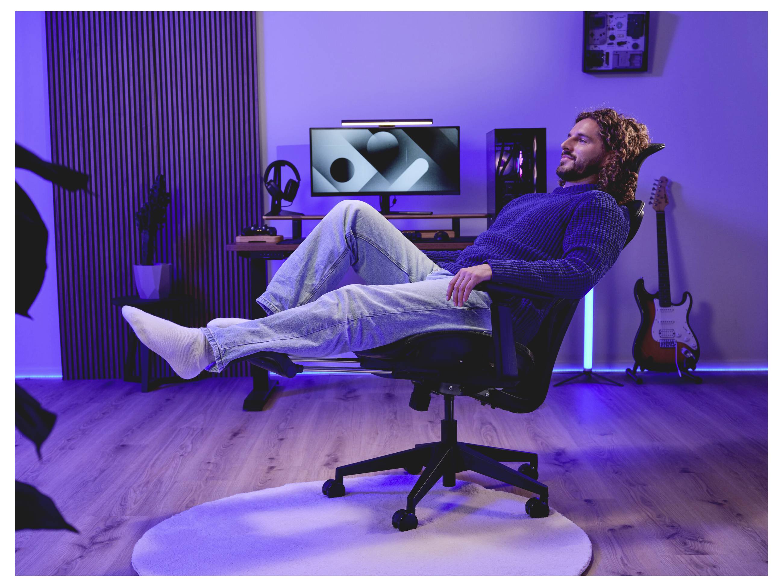 Ein Mann lehnt sich in einem Bürostuhl in einem violett beleuchteten Raum zurück, mit einem Desktop-Computer und einer Gitarre im Hintergrund.
