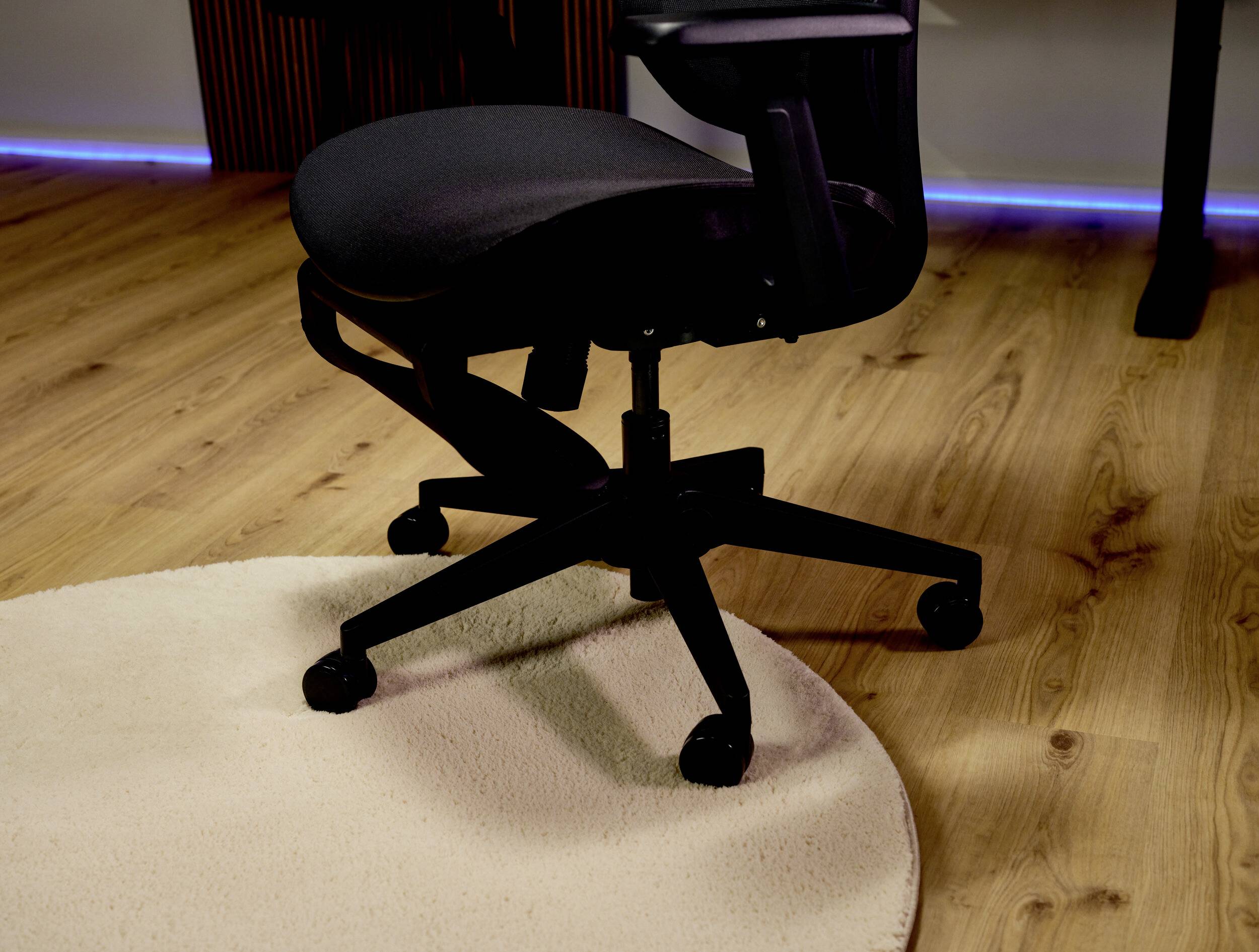 Schwarzer Drehbürostuhl auf einem Holzboden neben einem beigen Teppich, mit blauer LED-Beleuchtung entlang der Wand.