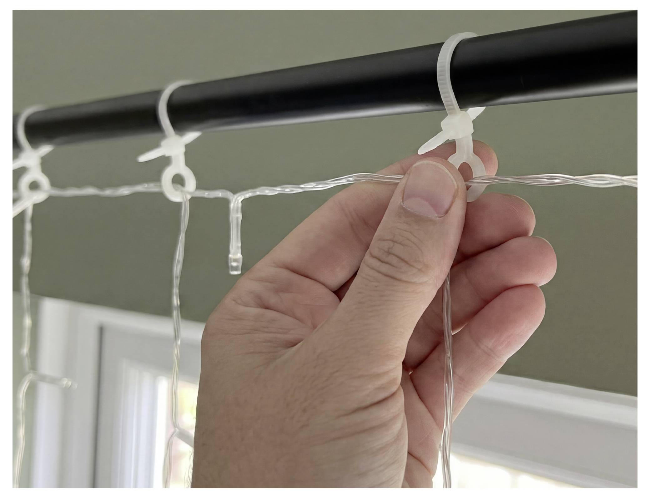Hand befestigt einen transparenten Schnurlichtklemmer an einer schwarzen Gardinenstange über einem Fenster.