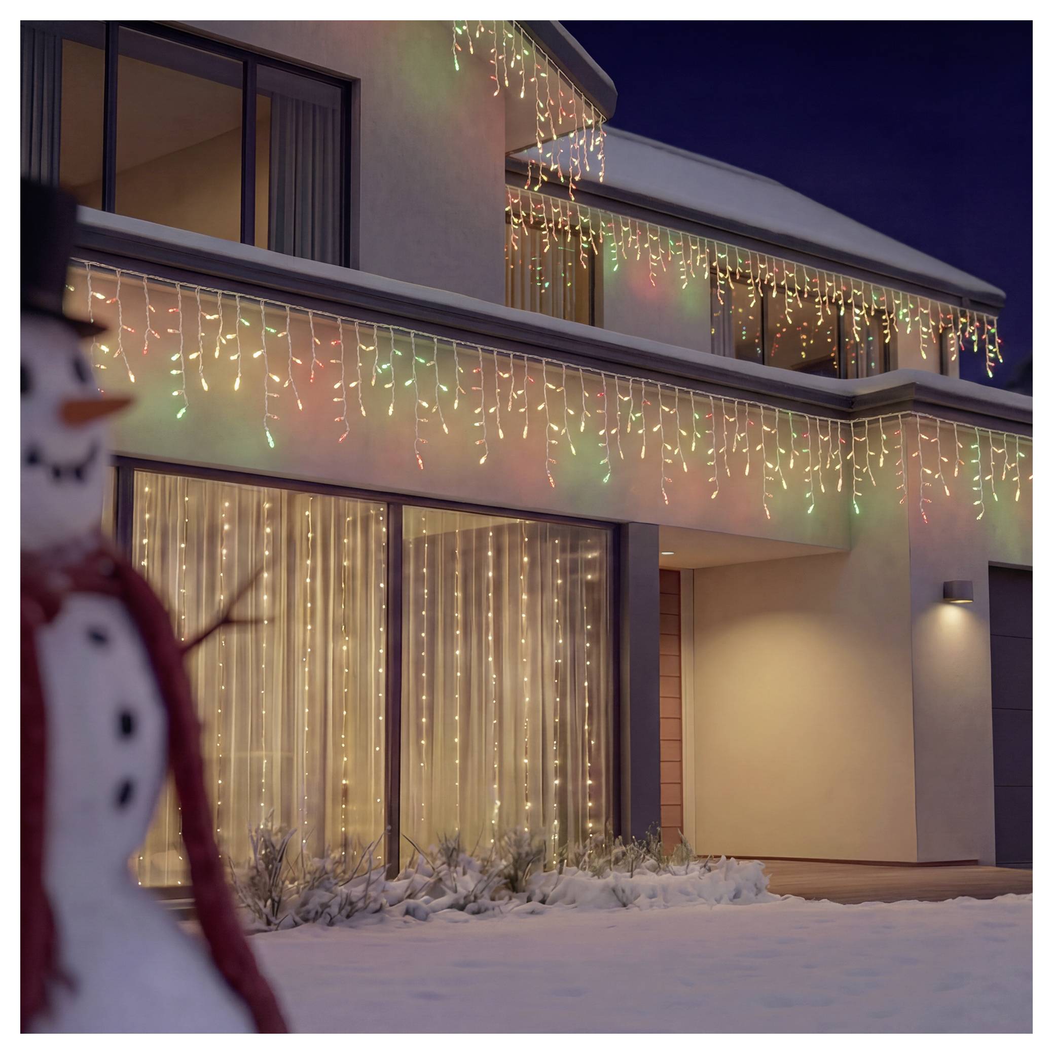 Schneebedecktes modernes Haus in der Nacht, geschmückt mit mehrfarbigen Weihnachtslichterketten, mit einem unscharfen Schneemann im Vordergrund.