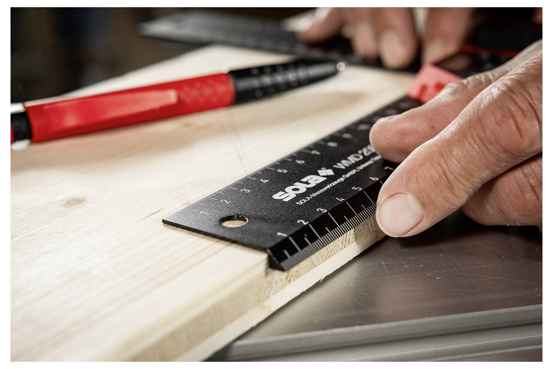 Nahaufnahme einer Person, die ein Holzbrett mit einem schwarzen Lineal misst, während ein rotes Handwerkzeug in der Nähe auf einer Arbeitsfläche liegt.